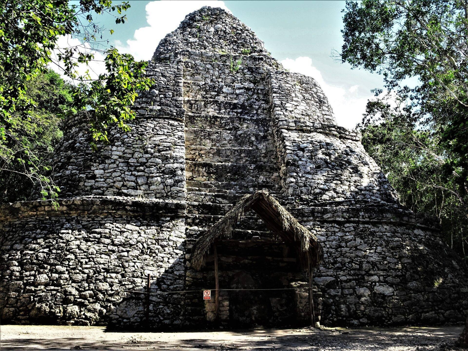 Steinerne Maya-Ruine mit zentraler Pyramide, umgeben von tropischer Vegetation und einem Strohdach am Eingang
