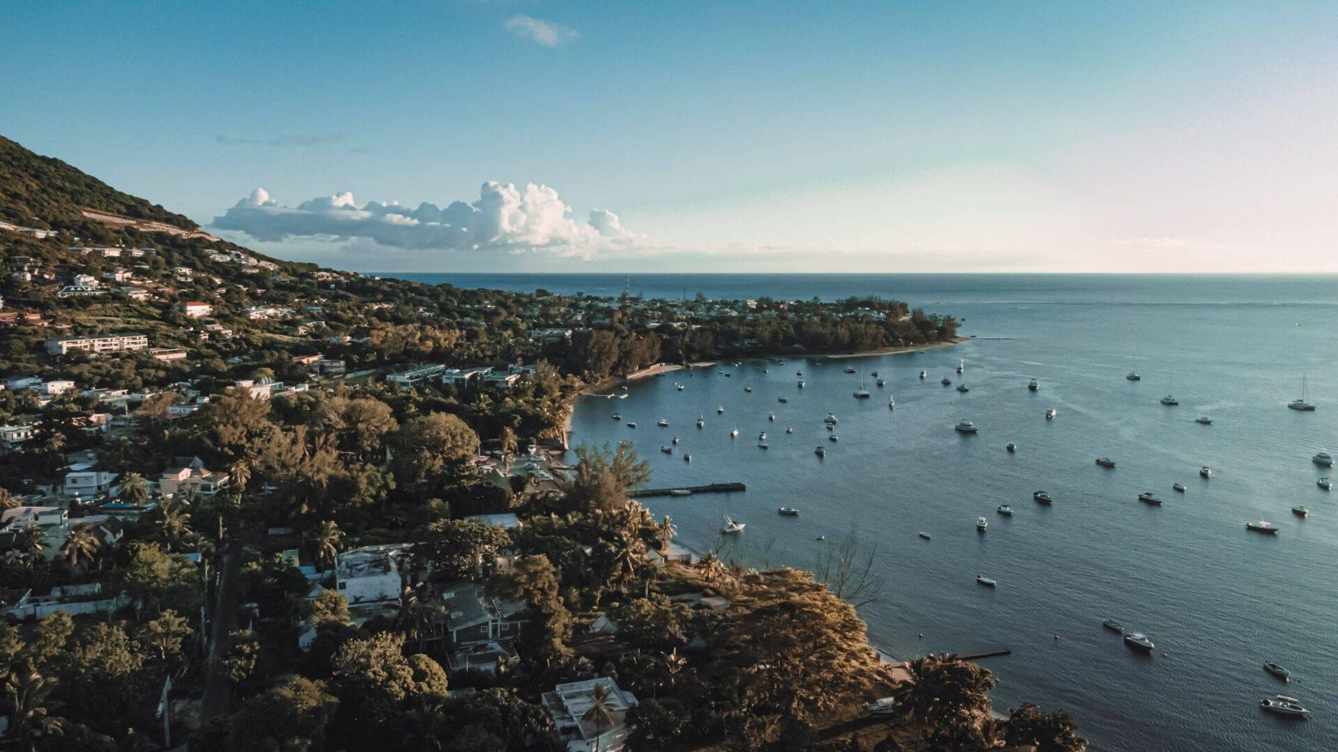 Una bahía costera tropical con casas dispersas sobre colinas boscosas, veleros fondeados y cielo despejado