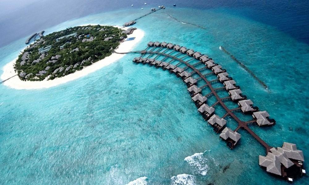 Vista aerea di un resort tropicale con bungalow sull’acqua, isola verdeggiante e laguna turchese alle Maldive