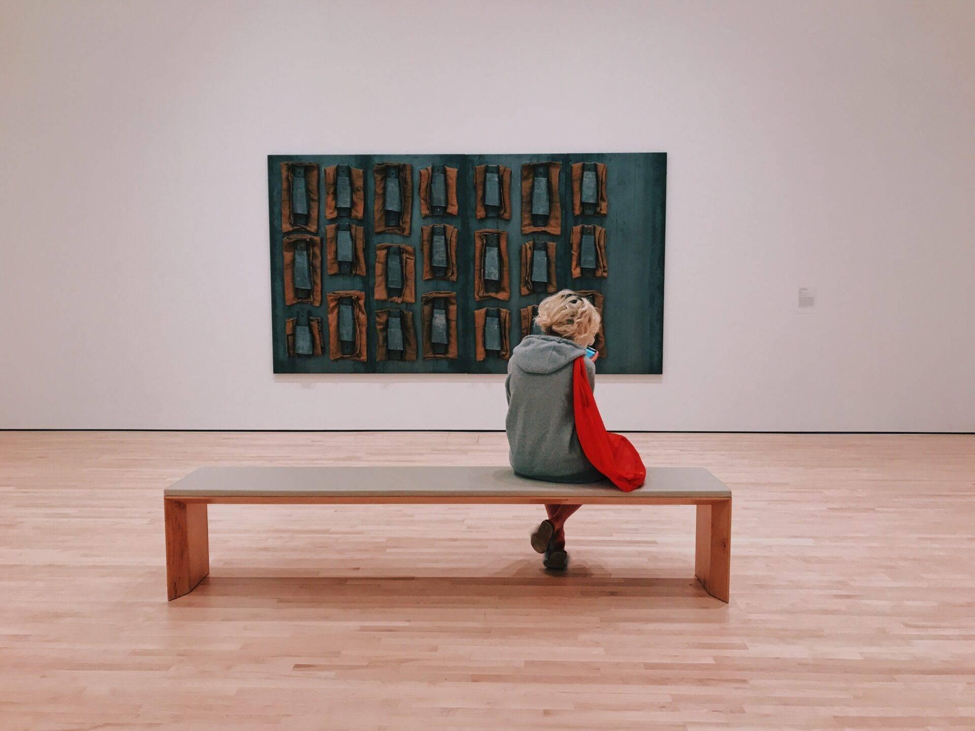 Eine Person sitzt auf einer Holzbank und betrachtet ein abstraktes Gemälde in Blau- und Orangetönen in einer Kunstgalerie
