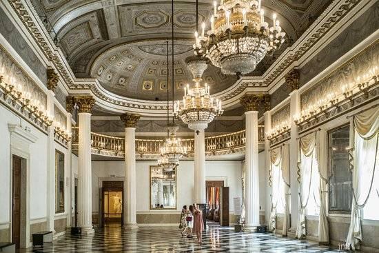Grande sala di palazzo con colonne bianche, lampadari di cristallo e soffitto ornamentale con decorazioni dorate