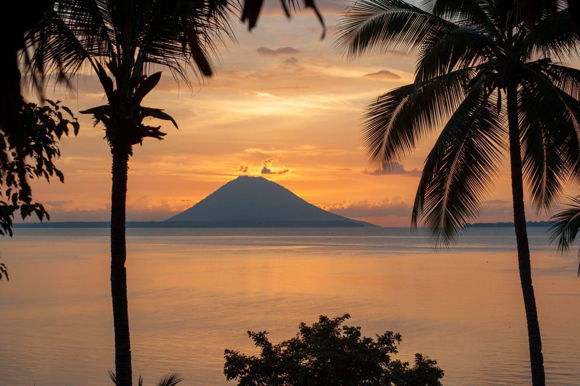 Tramonto tropicale su un vulcano in mare incorniciato da palme in controluce