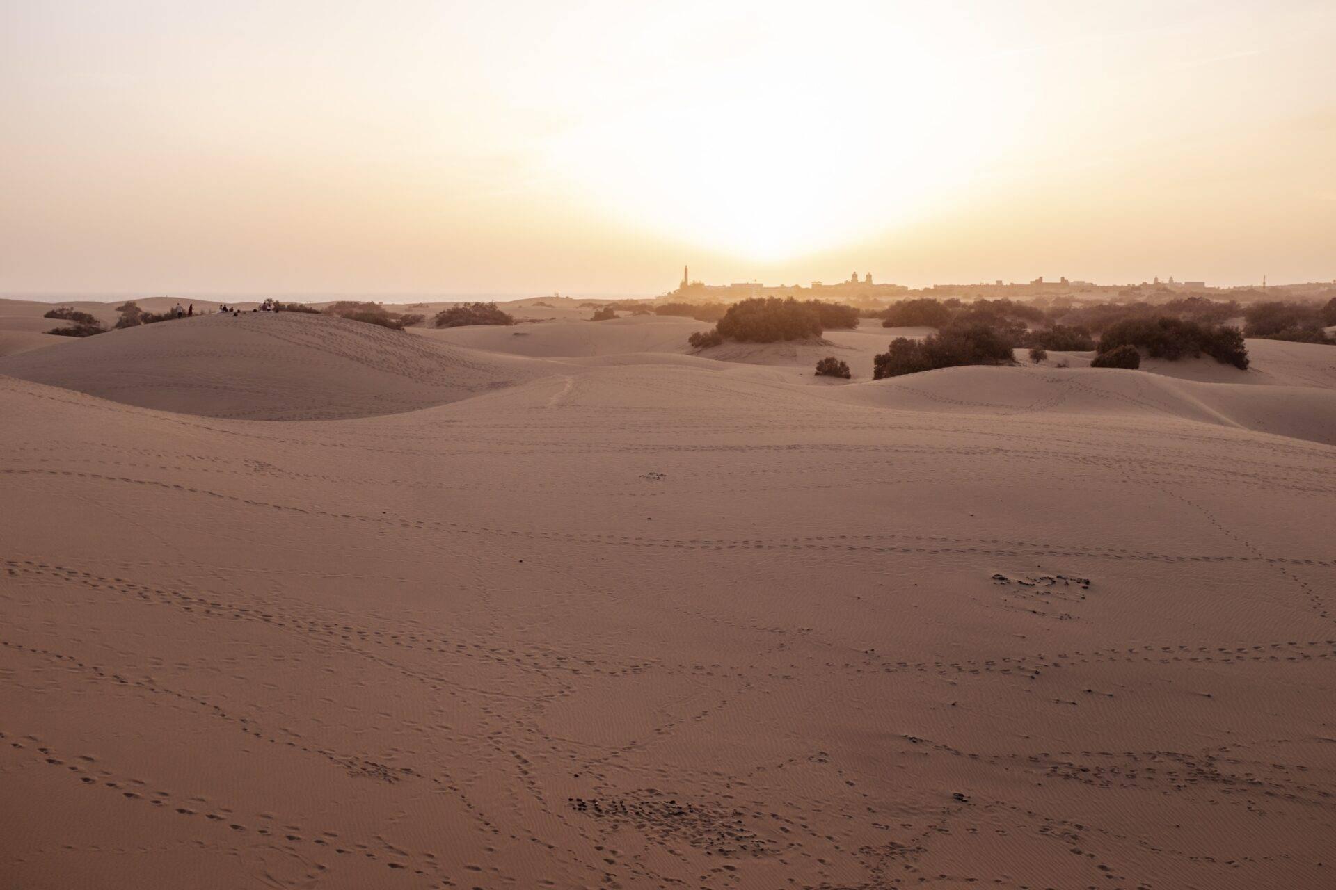 Paesaggio desertico con dune ondulate e un villaggio arroccato illuminato all’orizzonte al tramonto