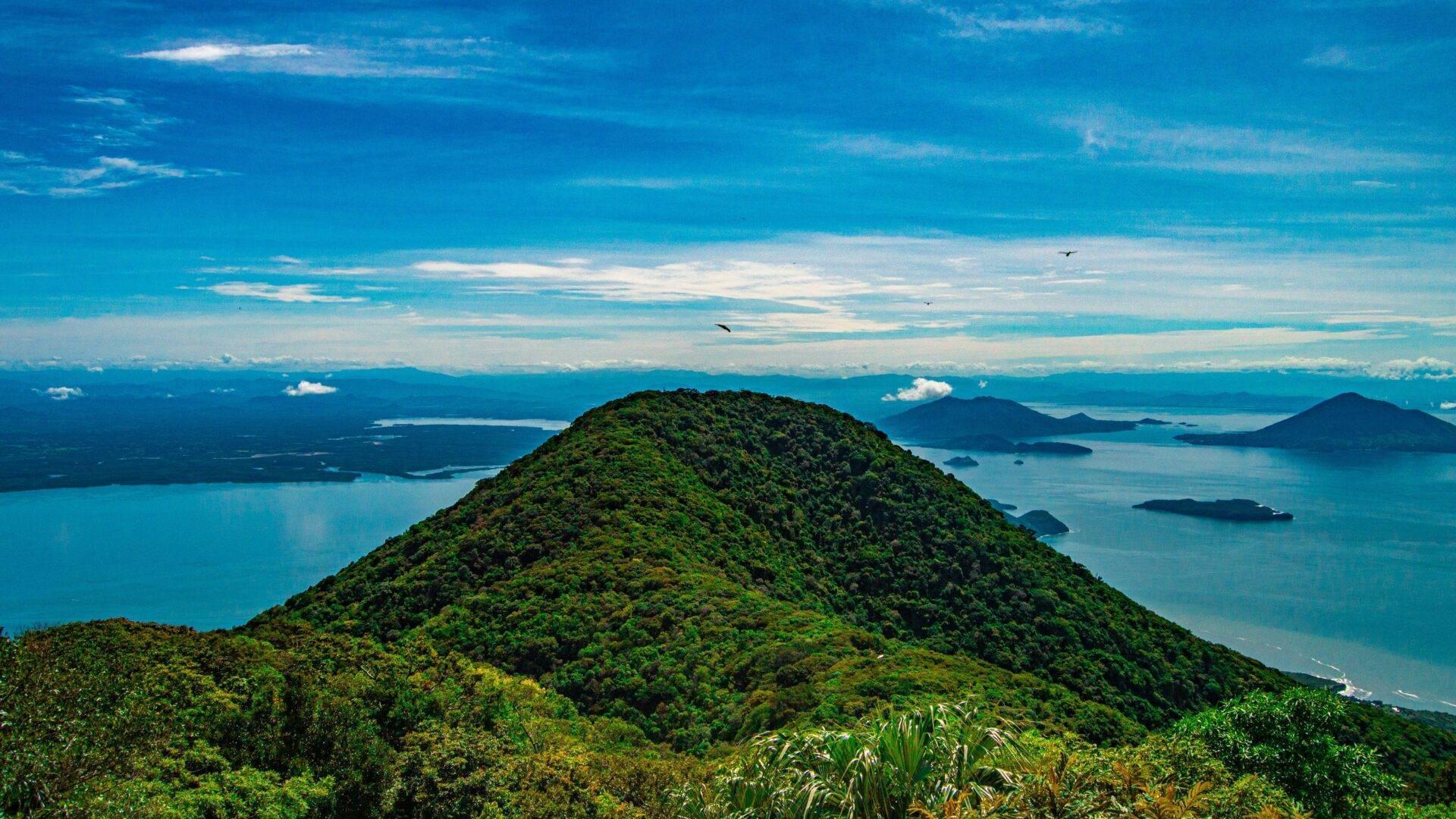 Vista aerea di un’isola densamente boscosa circondata da un mare turchese con isolotti e montagne sullo sfondo