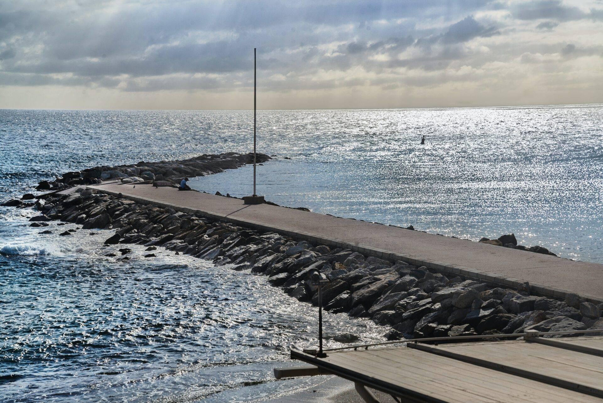 Un molo in cemento con un palo si protende verso il mare, costeggiato da rocce, con tempo nuvoloso