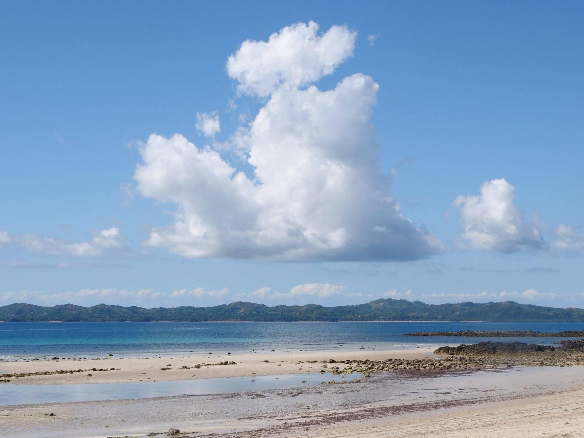 Una spiaggia di sabbia bianca con laguna turchese, montagne boscose sullo sfondo e cielo blu nuvoloso