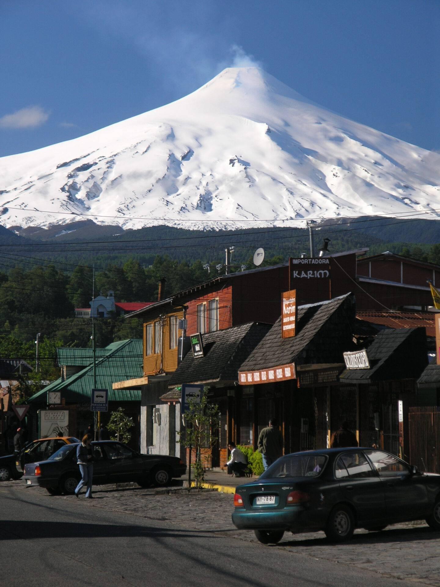 Una calle comercial con pequeños comercios de madera frente a un majestuoso volcán nevado bajo un cielo azul
