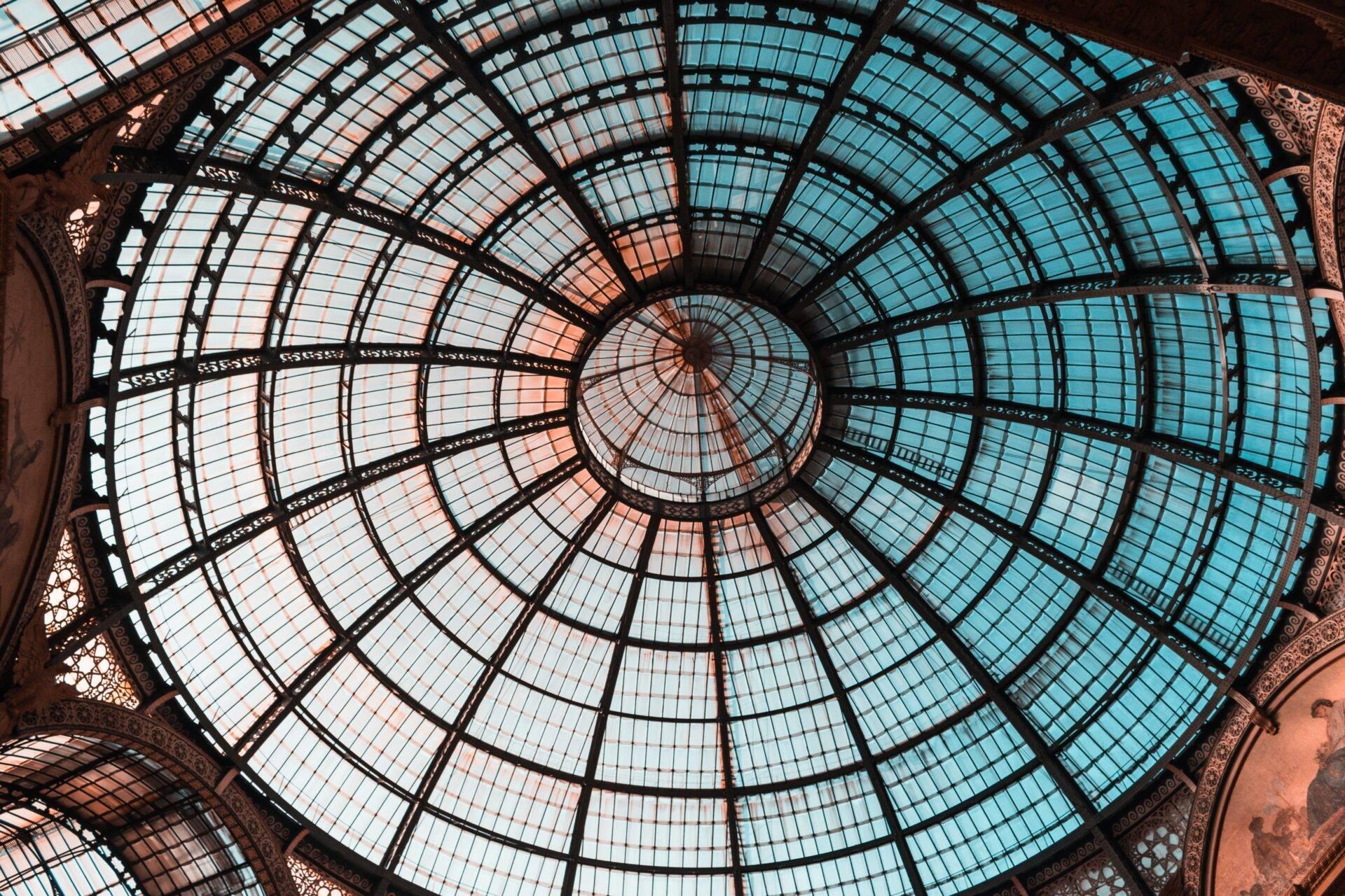 Overhead view of a domed architectural glass ceiling with geometric panes, ornamental metalwork and natural light flooding through