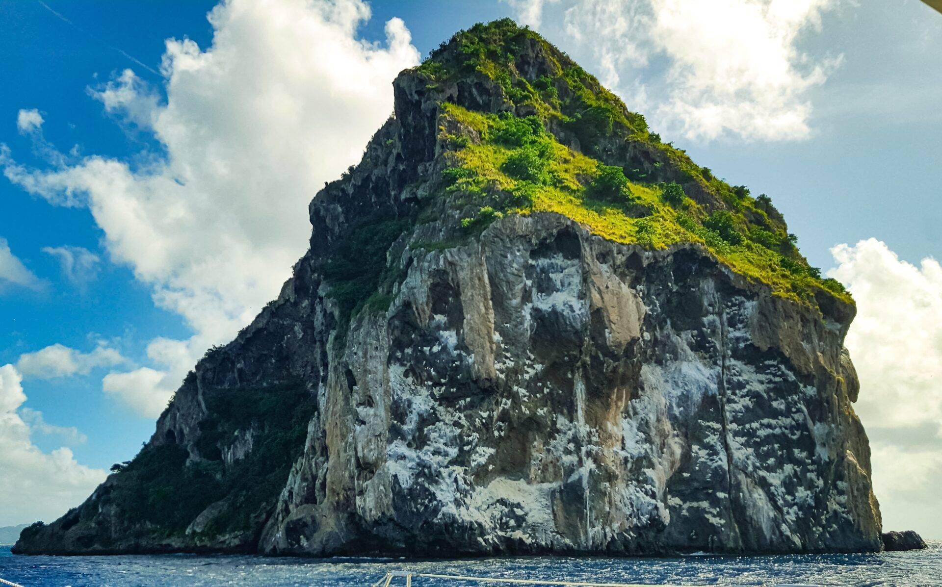 Ein steiler, begrünter Kalkfelsen erhebt sich aus dem türkisfarbenen Meer unter wolkenlosem blauem Himmel