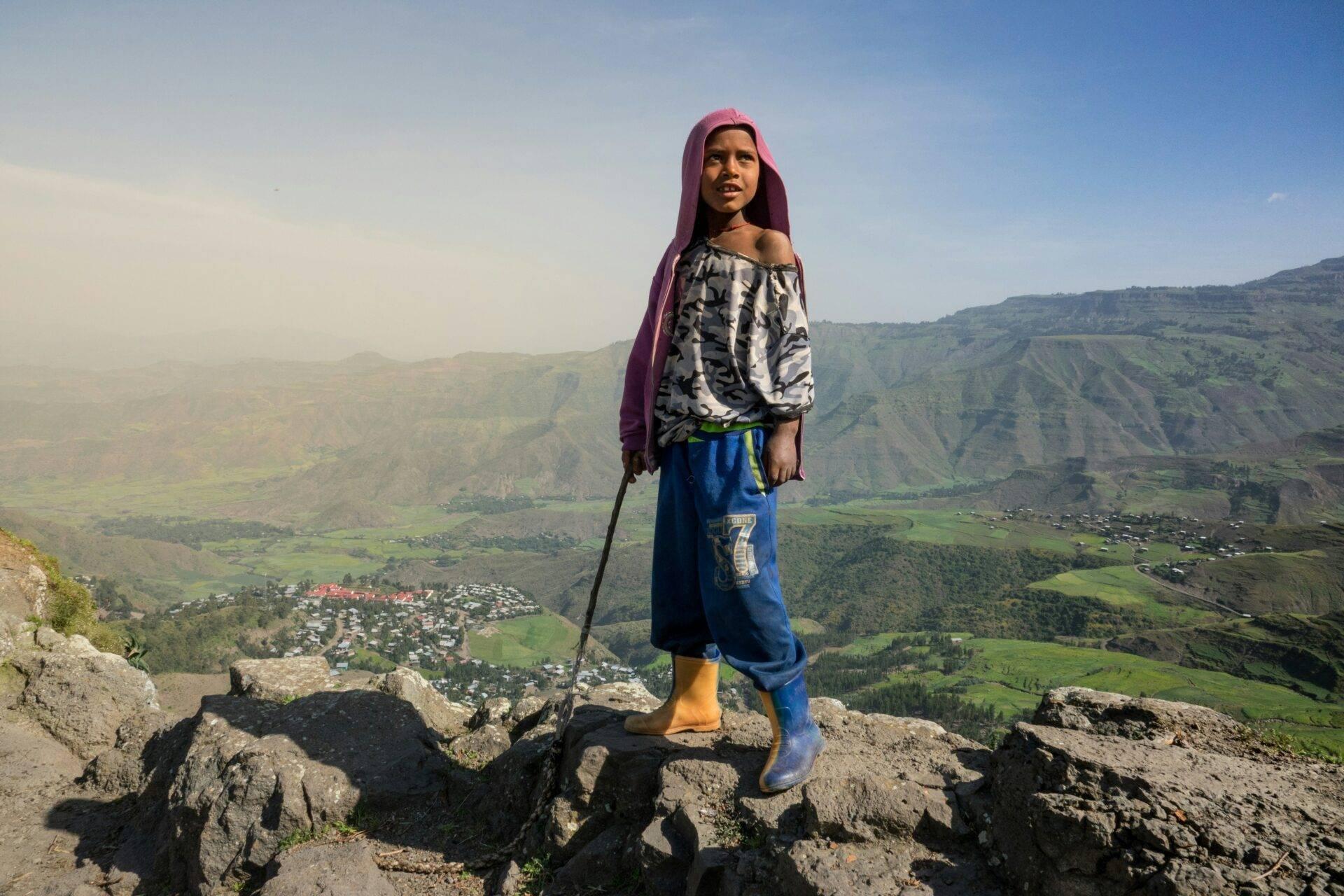 A young girl in traditional dress poses on rocky high ground above a lush green mountain valley