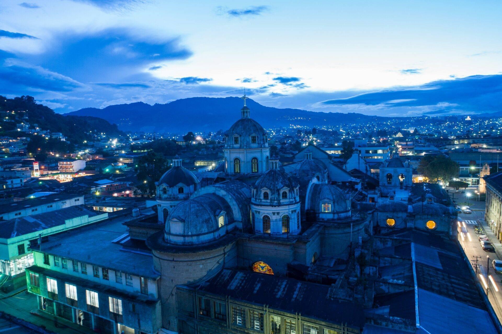 Luftaufnahme einer kolonialen Kathedrale mit blauen Kuppeln, in der Abenddämmerung beleuchtet, umgeben von Bergen und Häusern
