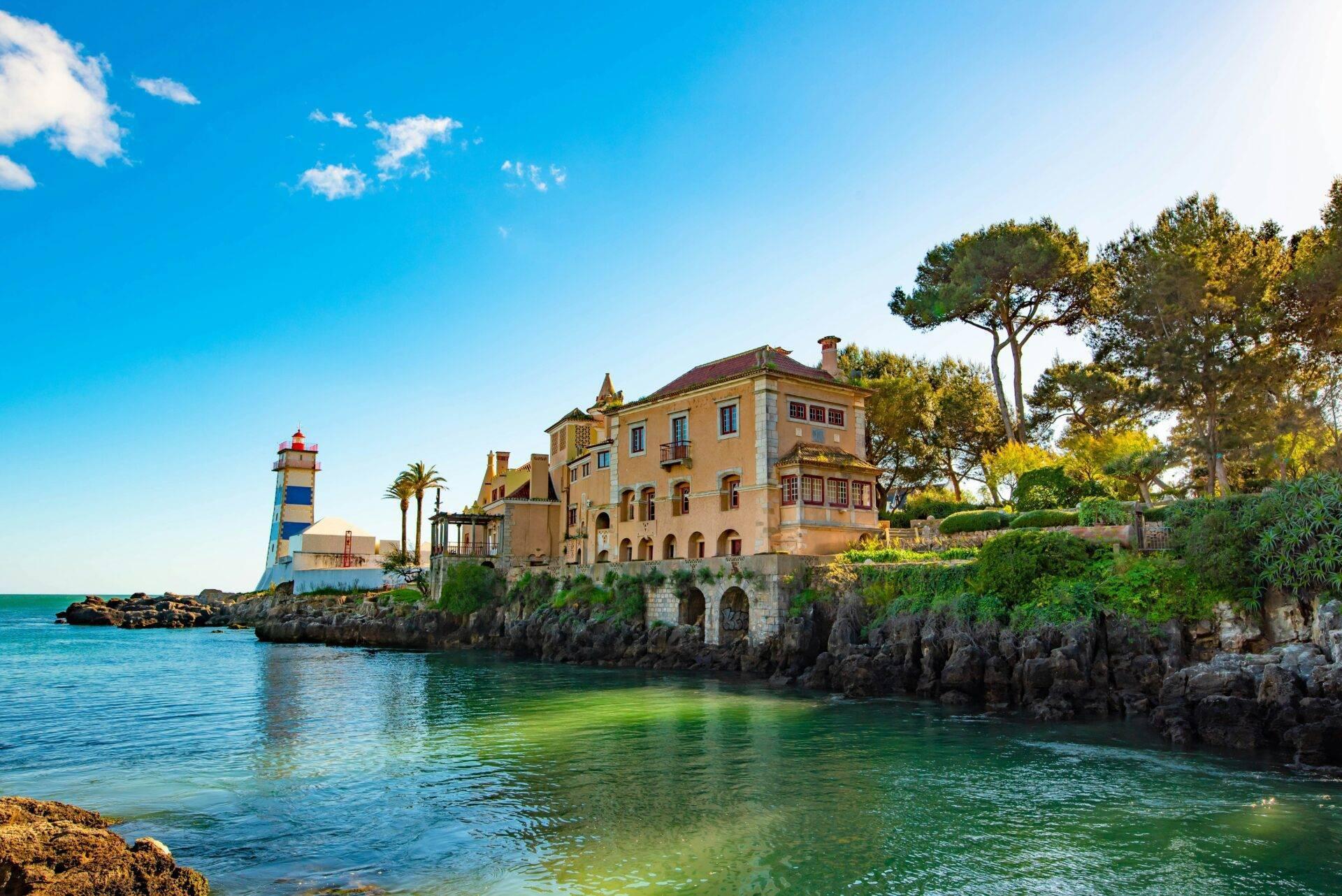 Ein ockerfarbener Palast auf Küstenfelsen mit einem blau-weißen Leuchtturm, der über eine türkisblaue Bucht ragt