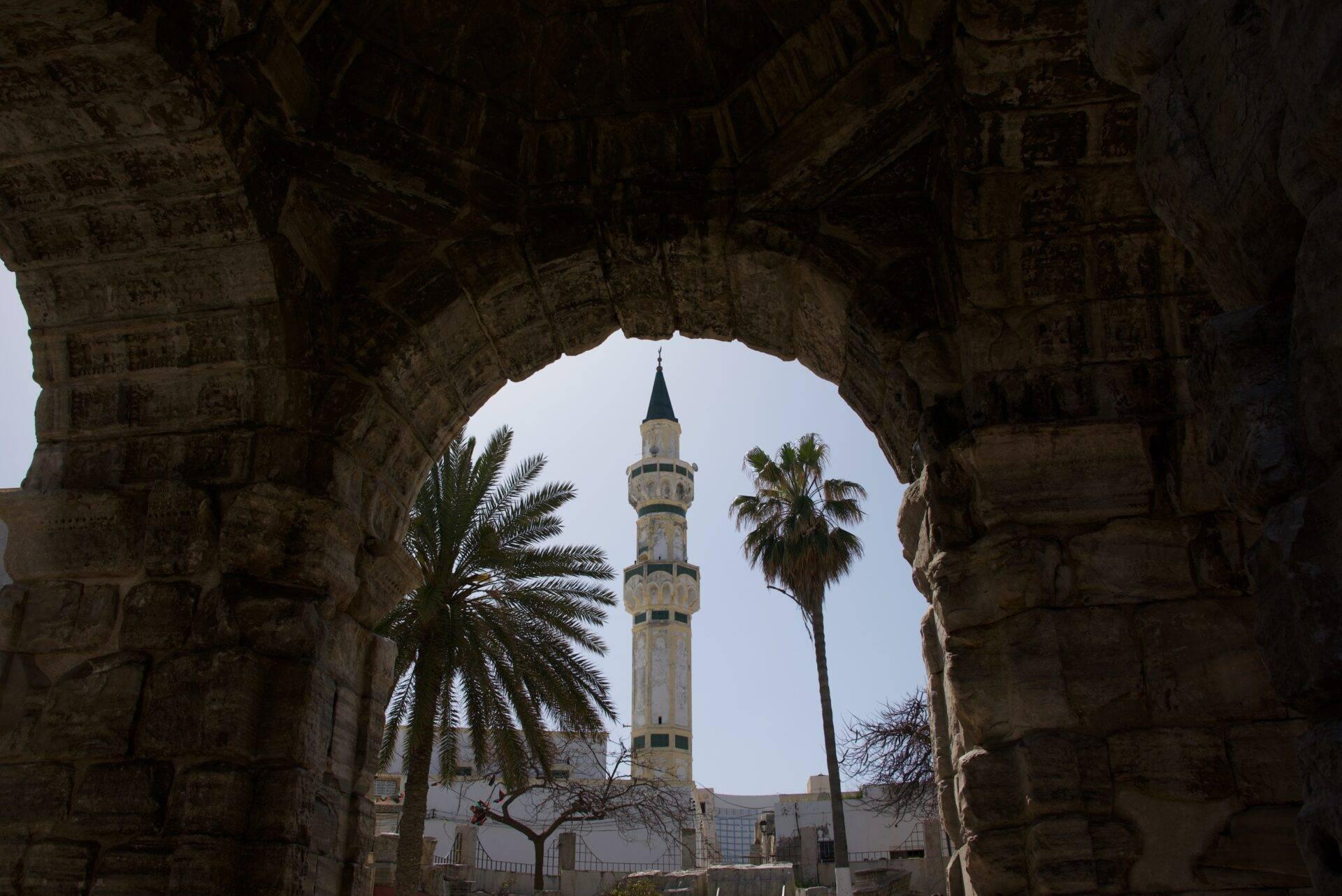 Vista a través de un arco de piedra de un minarete blanco con balcones azules y palmeras