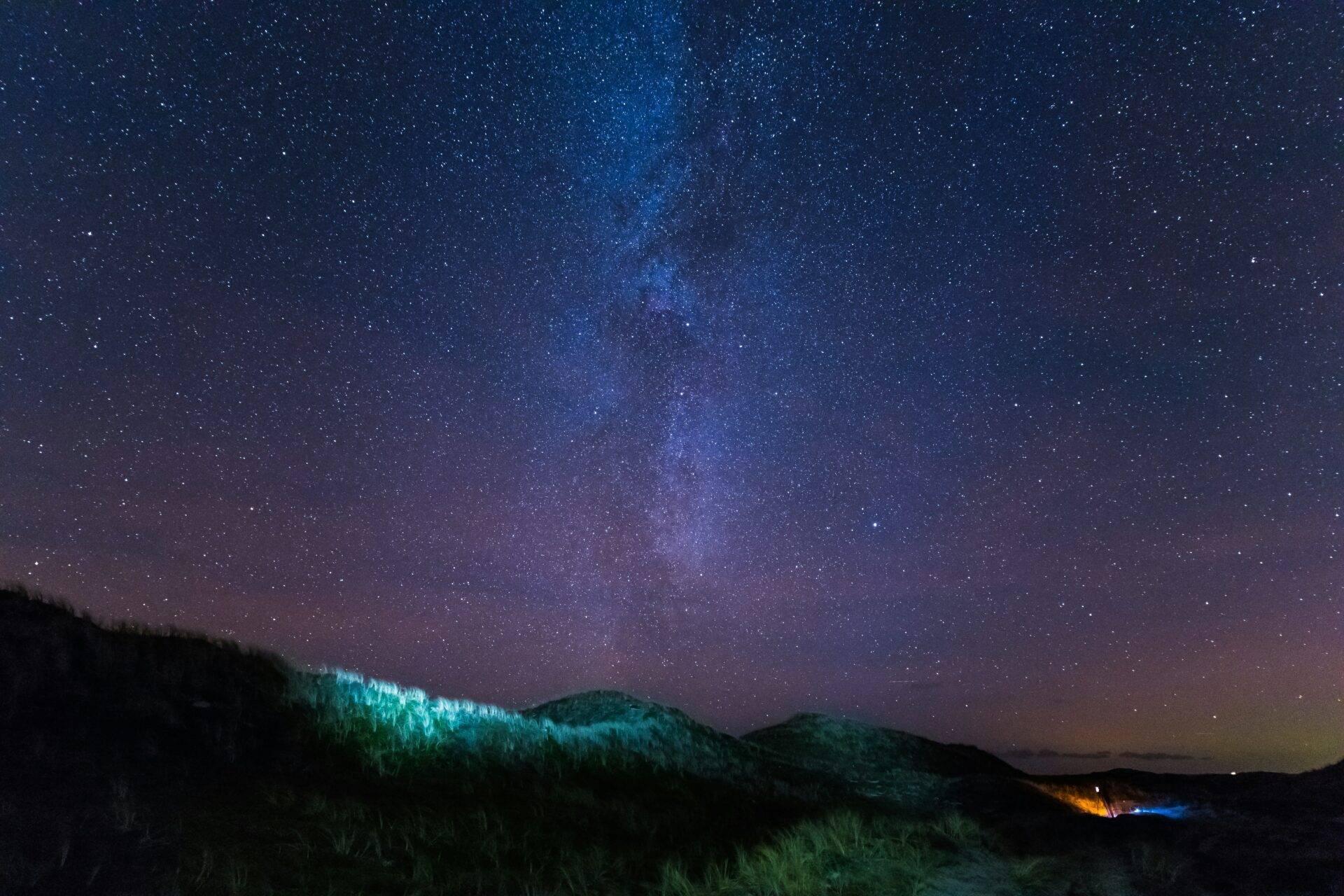 Montagne boscose sotto un cielo stellato con la Via Lattea visibile e alcune luci in lontananza