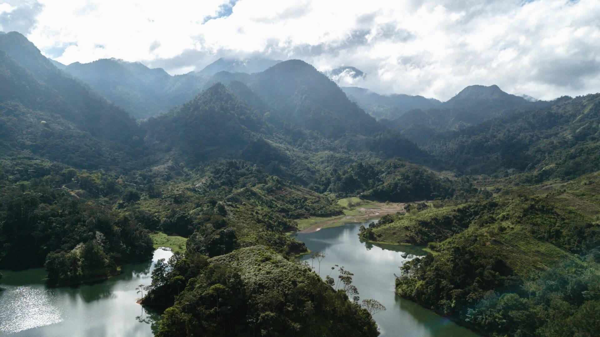 Valle verdeggiante circondata da montagne boscose con fiumi e laghi che riflettono il cielo nuvoloso