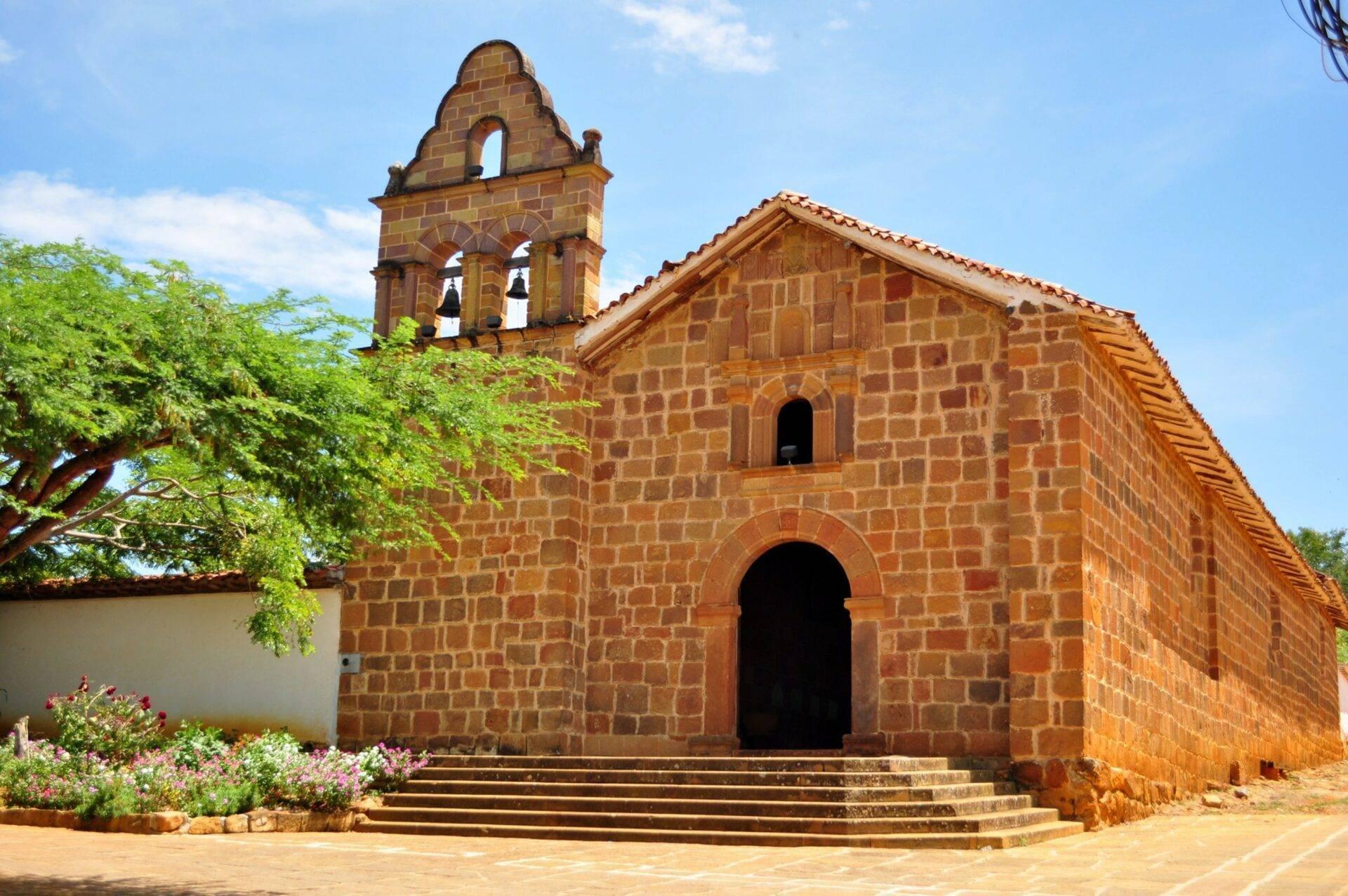 Iglesia colonial de ladrillo rojo con campanario bajo un cielo azul, rodeada de vegetación tropical