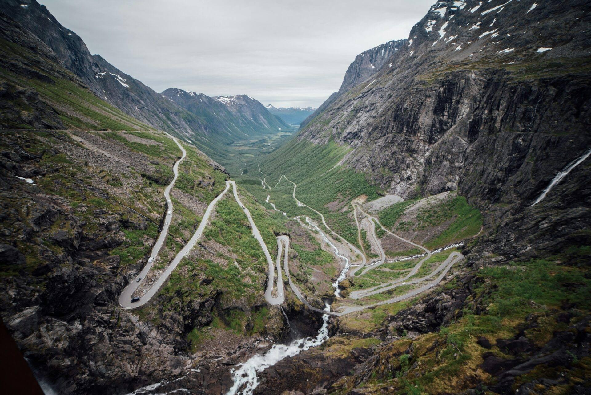 Eine kurvige Straße führt durch einen alpinen Pass mit grünem Tal und Wasserfall im Hintergrund