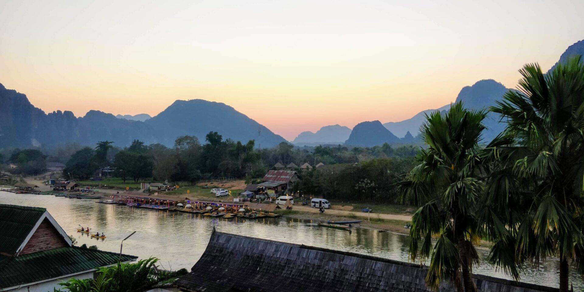 Vista panoramica di una valle fluviale in Asia sud-orientale al tramonto, con montagne calcaree, palme e villaggio tradizionale