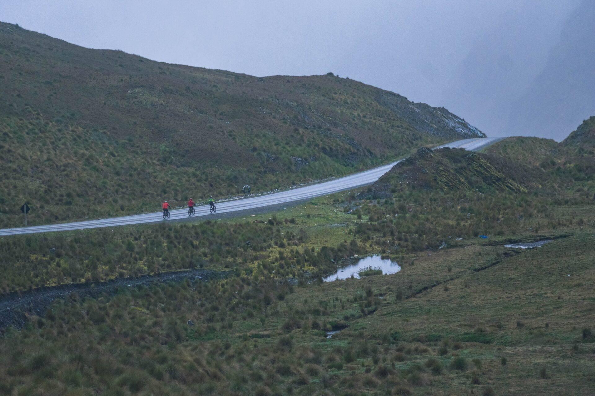 Strada asfaltata sinuosa che attraversa montagne boscose con ciclisti e un paesaggio di tundra alpina