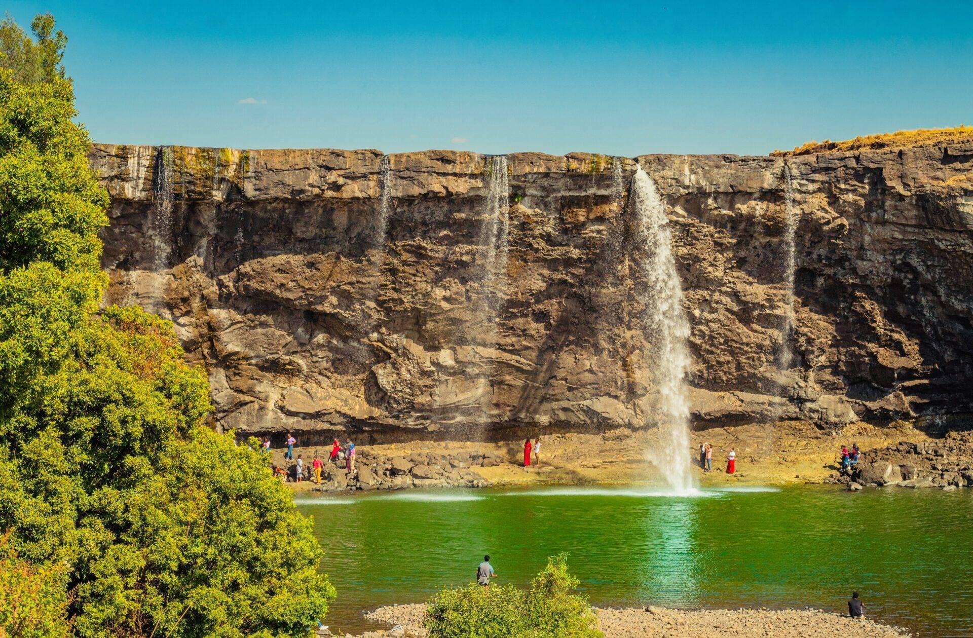 A spectacular waterfall cascading from a rocky cliff into a turquoise pool, surrounded by visitors and lush green vegetation
