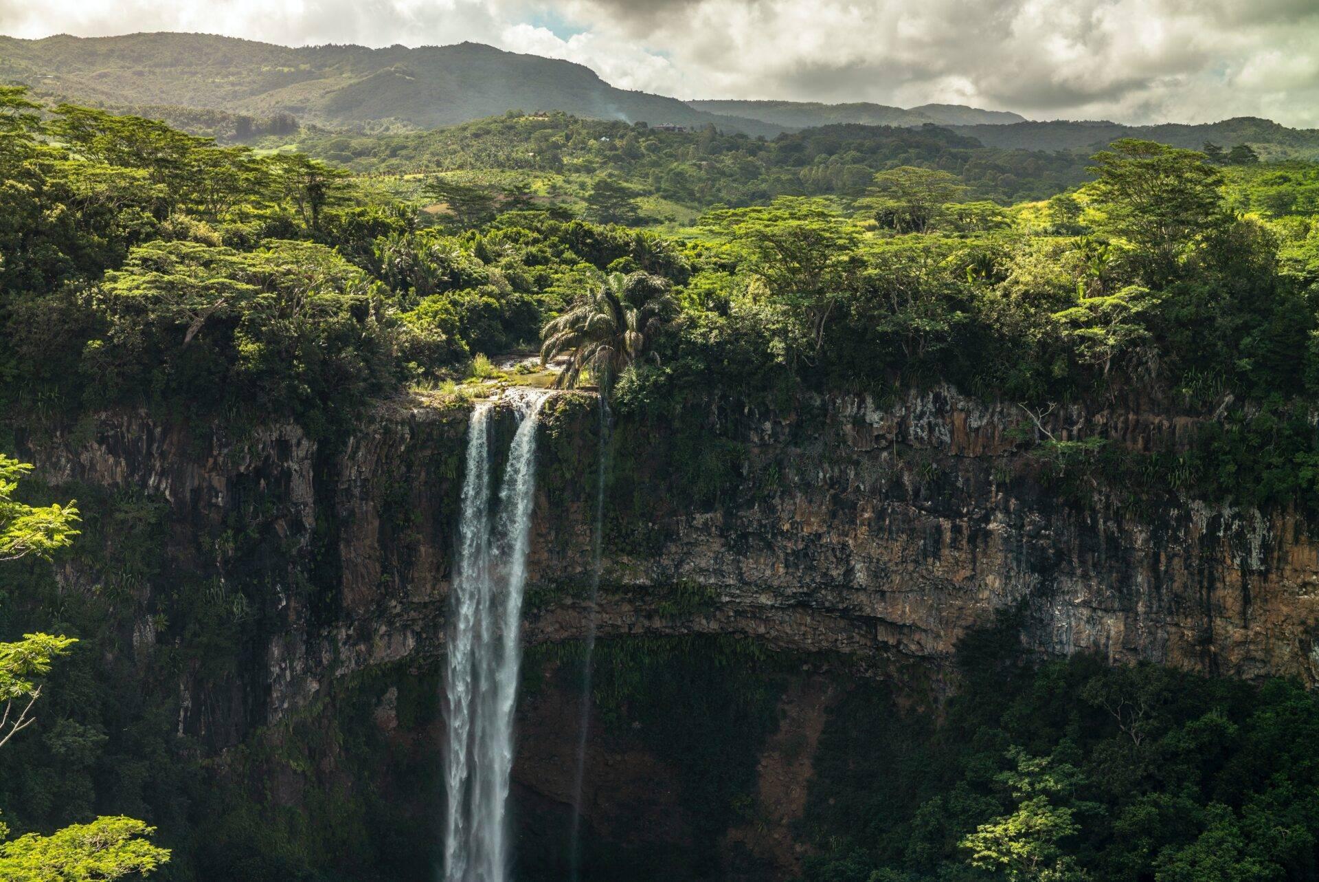 Una espectacular cascada cae desde un acantilado basáltico hacia un exuberante valle tropical rodeado de montañas boscosas