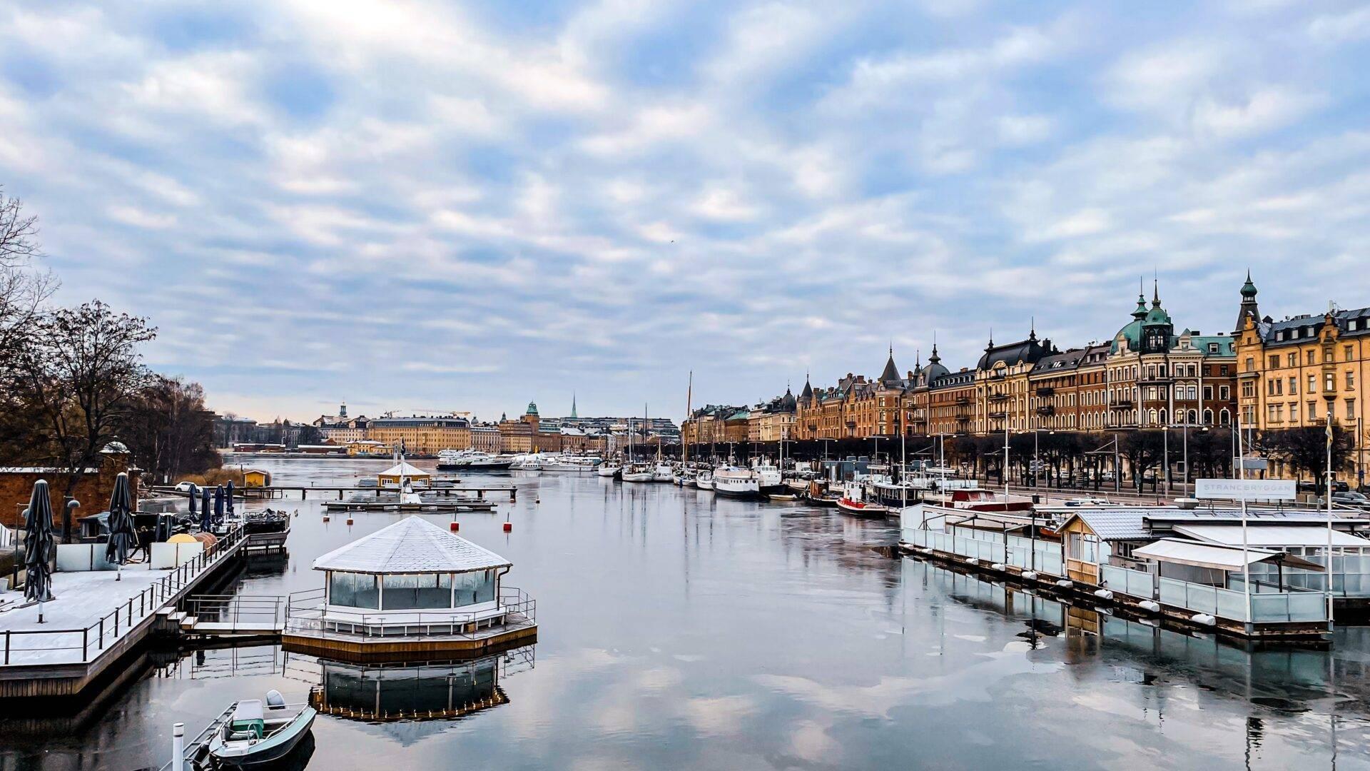 Winterlicher Blick auf einen skandinavischen Hafen mit festgemachten Booten, farbiger Architektur und gefrorenem Wasser, das den bewölkten Himmel spiegelt