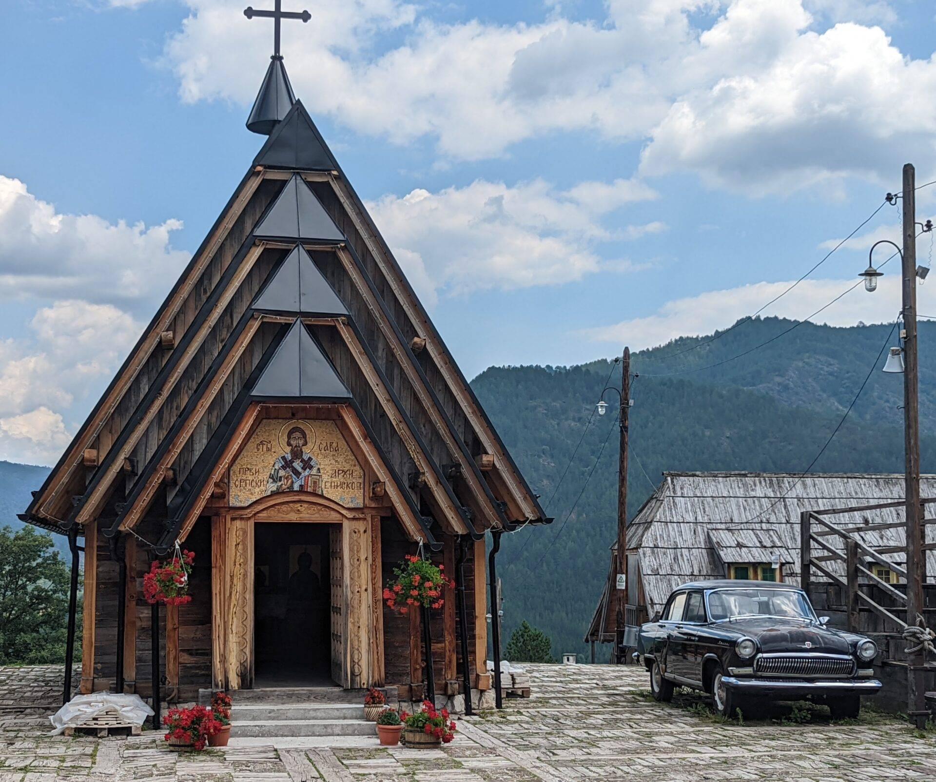 Eine kleine orthodoxe Holzkirche mit goldenem Kreuz, einem Oldtimer und bewaldeten Bergen im Hintergrund