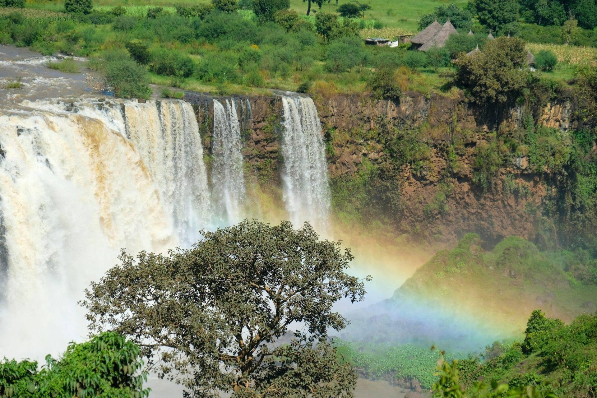 Spektakuläre kaskadenartige Wasserfälle über Felswände mit Regenbogen, üppiger Vegetation und traditionellen Häusern im Hintergrund