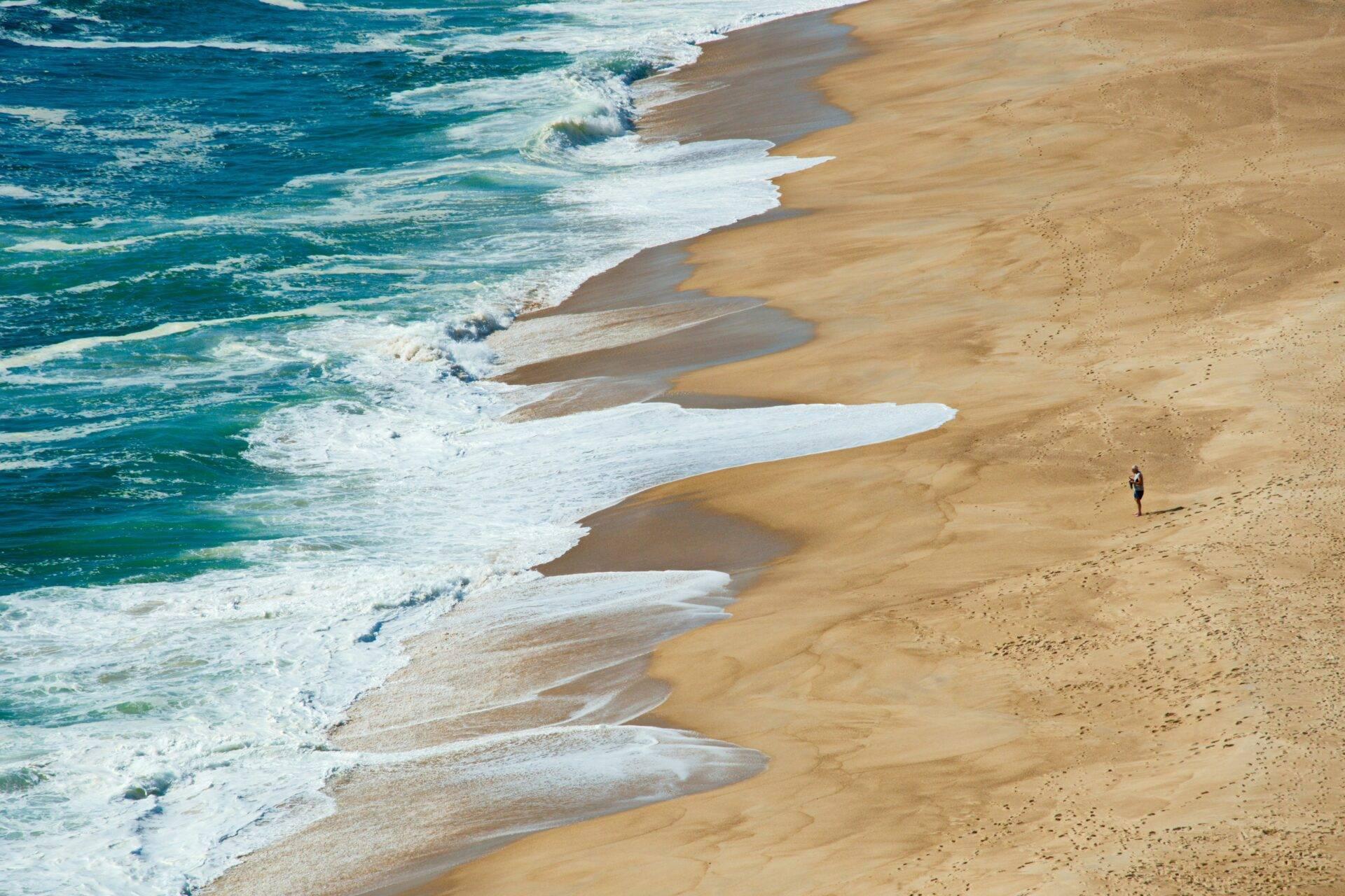 Luftaufnahme eines goldenen Sandstrands mit Brandungswellen und einem einzelnen Spaziergänger in Portugal