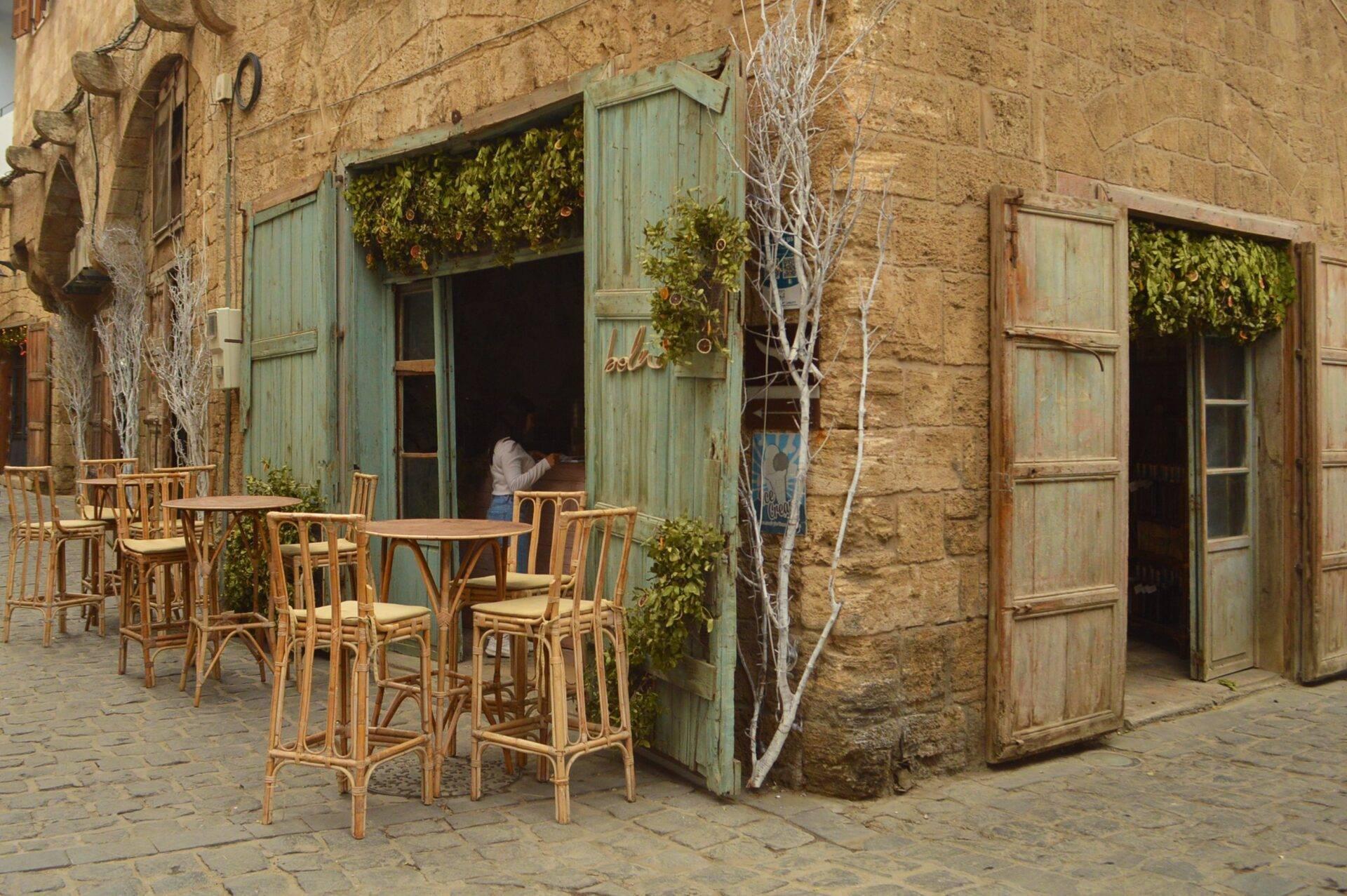 Terraza de café con sillas de mimbre frente a una fachada de piedra dorada y puertas verdes envejecidas