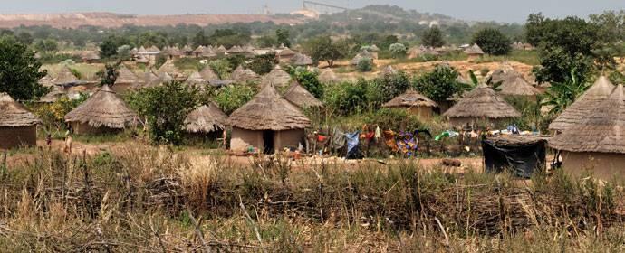 Villaggio tradizionale con capanne ai tetti di paglia, campi coltivati e abitanti in una zona rurale dell’Africa occidentale