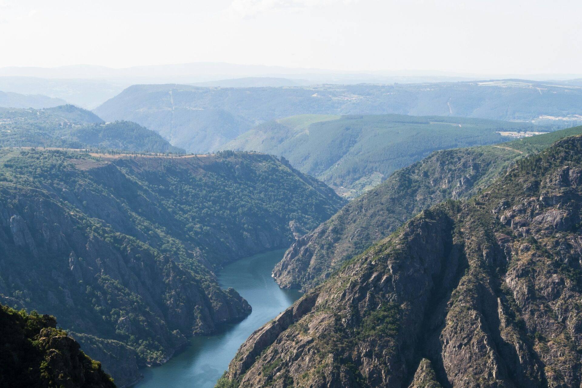 Tiefe Schlucht mit mäanderndem türkisblauem Fluss, umgeben von bewaldeten Felsen und fernen Bergen
