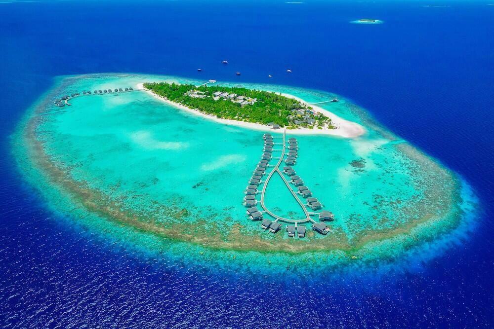 Vista aerea di un atollo tropicale con isola verdeggiante, bungalow sull’acqua e laguna turchese cristallina