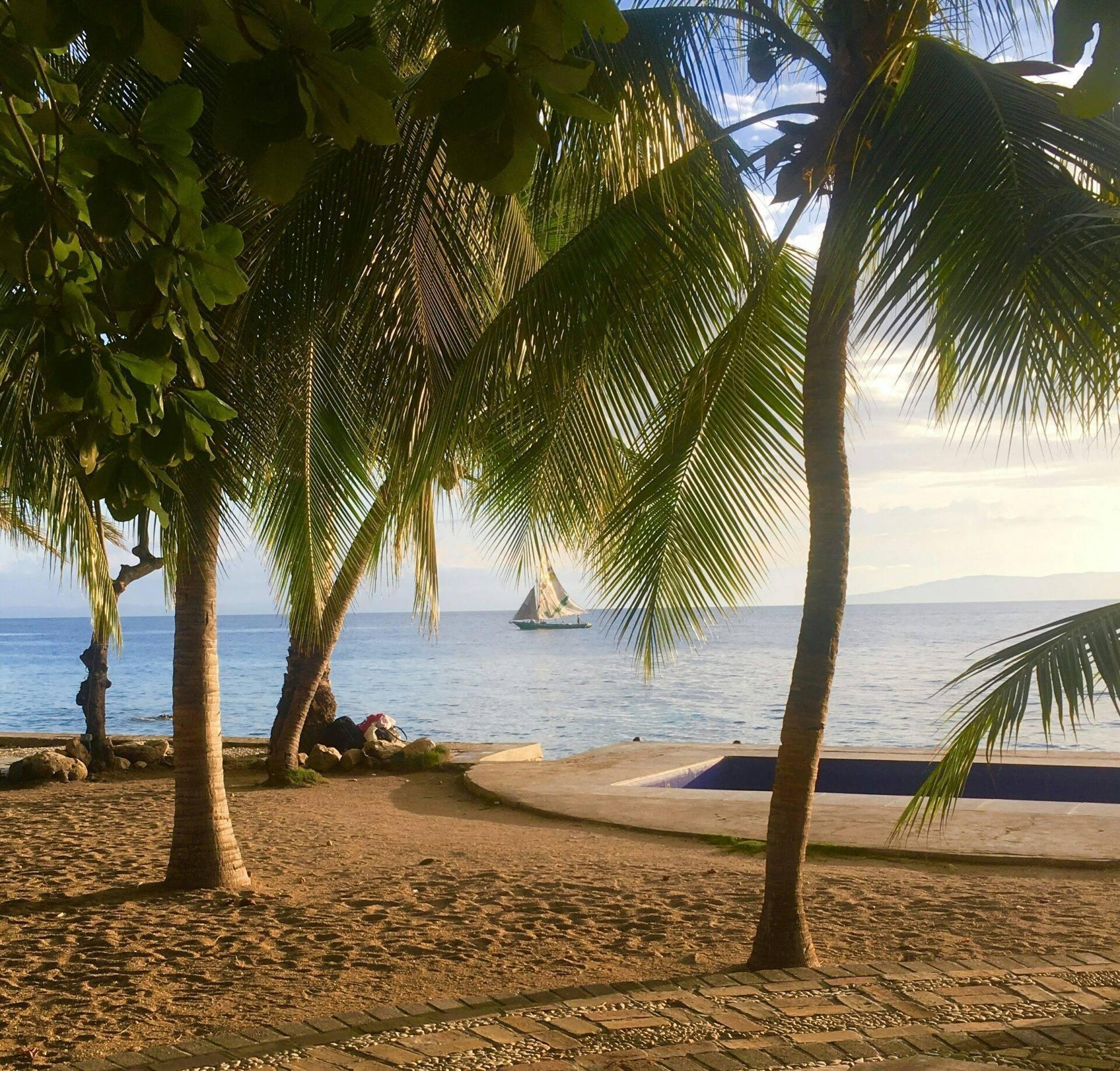 Playa tropical con palmeras, velero en el horizonte y piragua varada en la arena