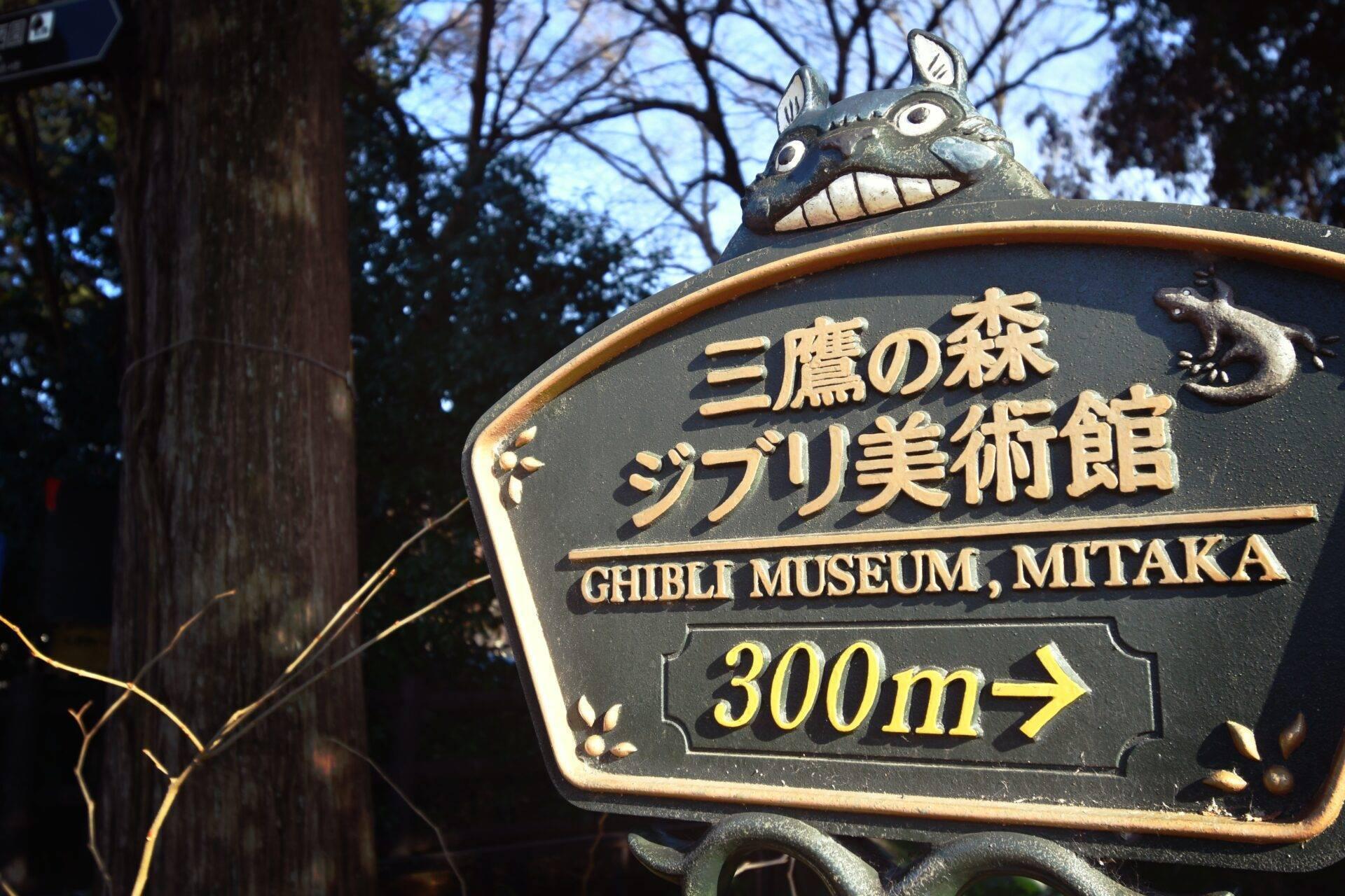 Wegweiserschild zum Ghibli-Museum Mitaka, verziert mit einer Fantasiegestalt, inmitten eines japanischen Waldes