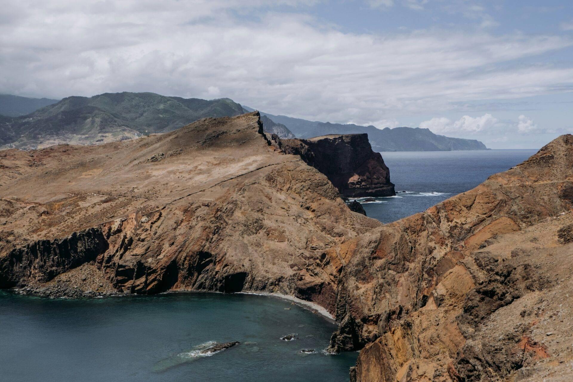 Arid coastal landscape with rocky cliffs, a small turquoise bay and volcanic islands in the background under a cloudy sky