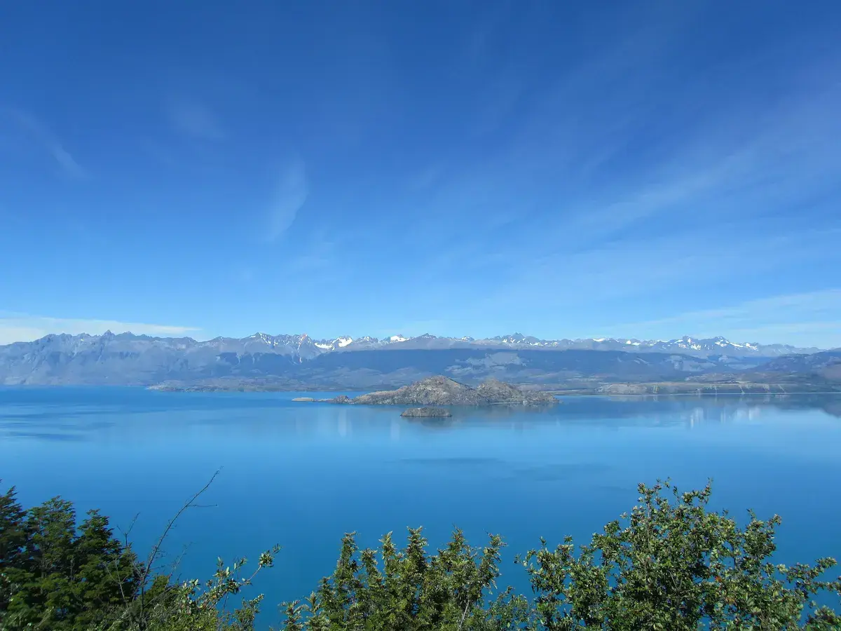 Lago alpino turquesa con islotes rocosos y una cadena montañosa nevada al fondo bajo cielo azul
