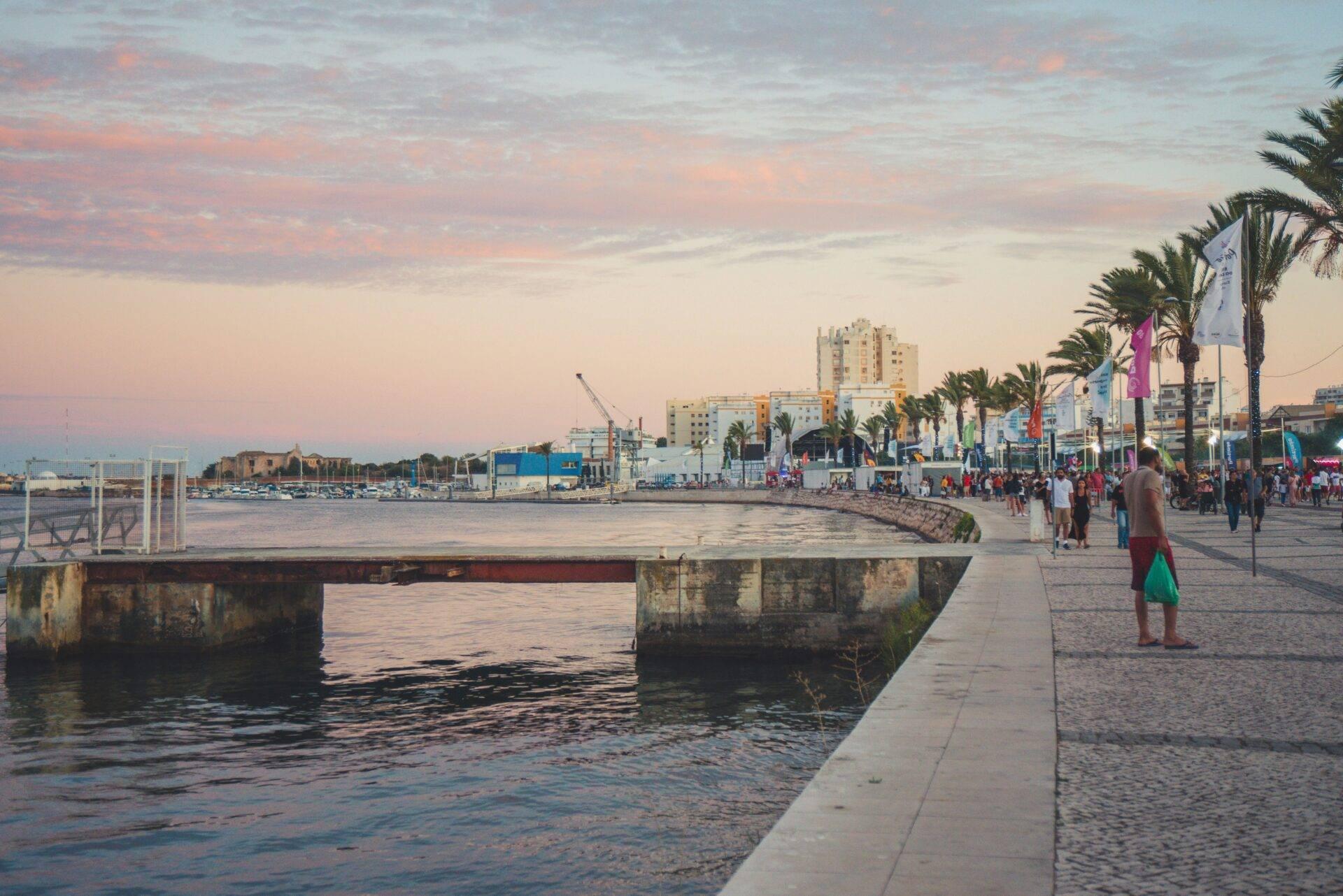 Eine belebte Küstenpromenade im Sonnenuntergang mit Palmen, moderner Architektur und einem Hafen im Hintergrund