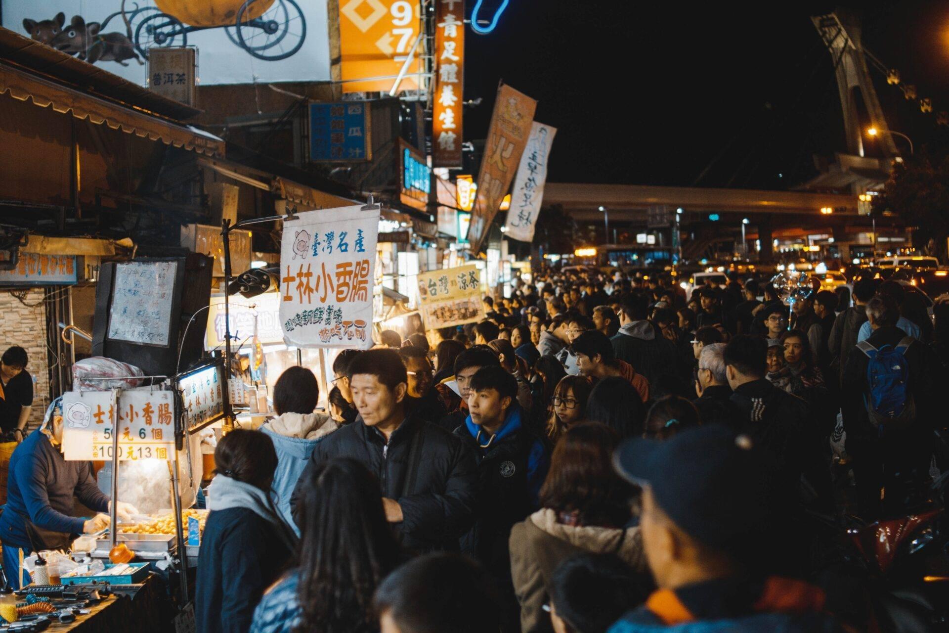 Una strada commerciale animata di notte con venditori di street food e una folla densa sotto insegne luminose in caratteri cinesi