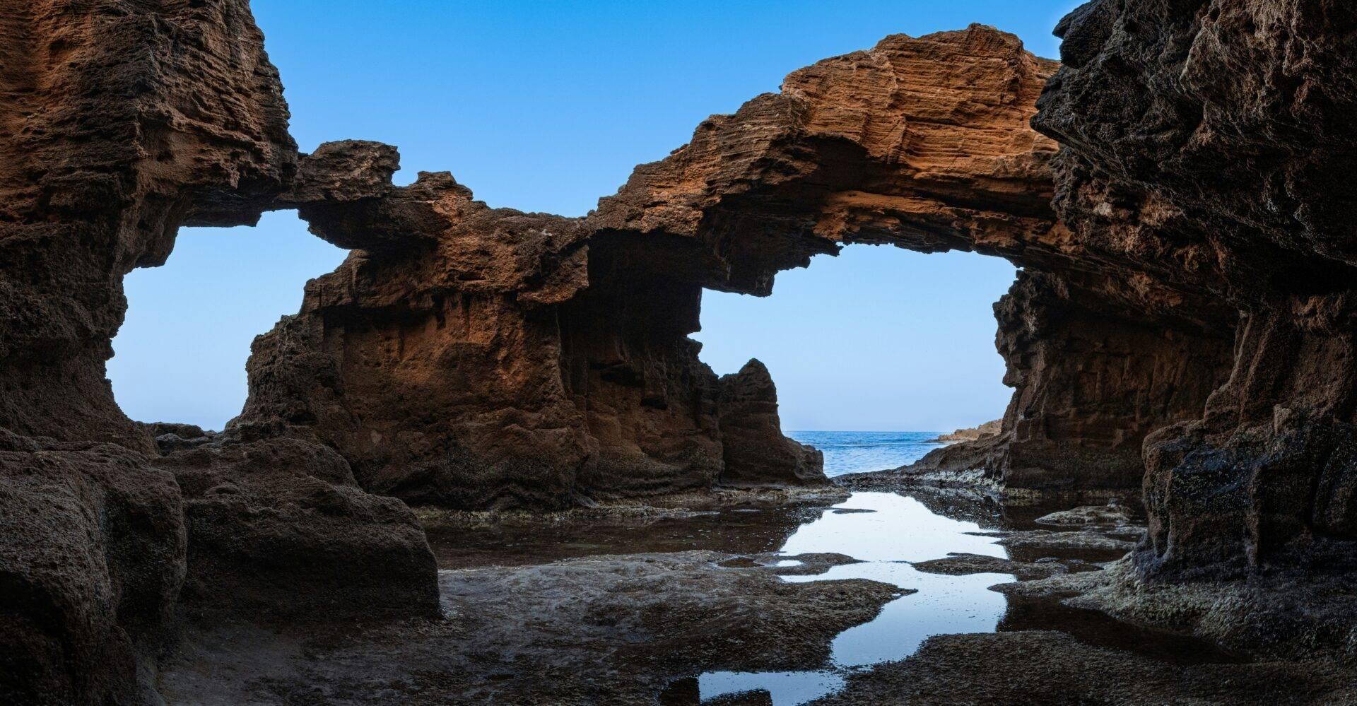 Ruines de pierre avec arches naturelles surplombant une plage rocheuse et des flaques d’eau réfléchissantes