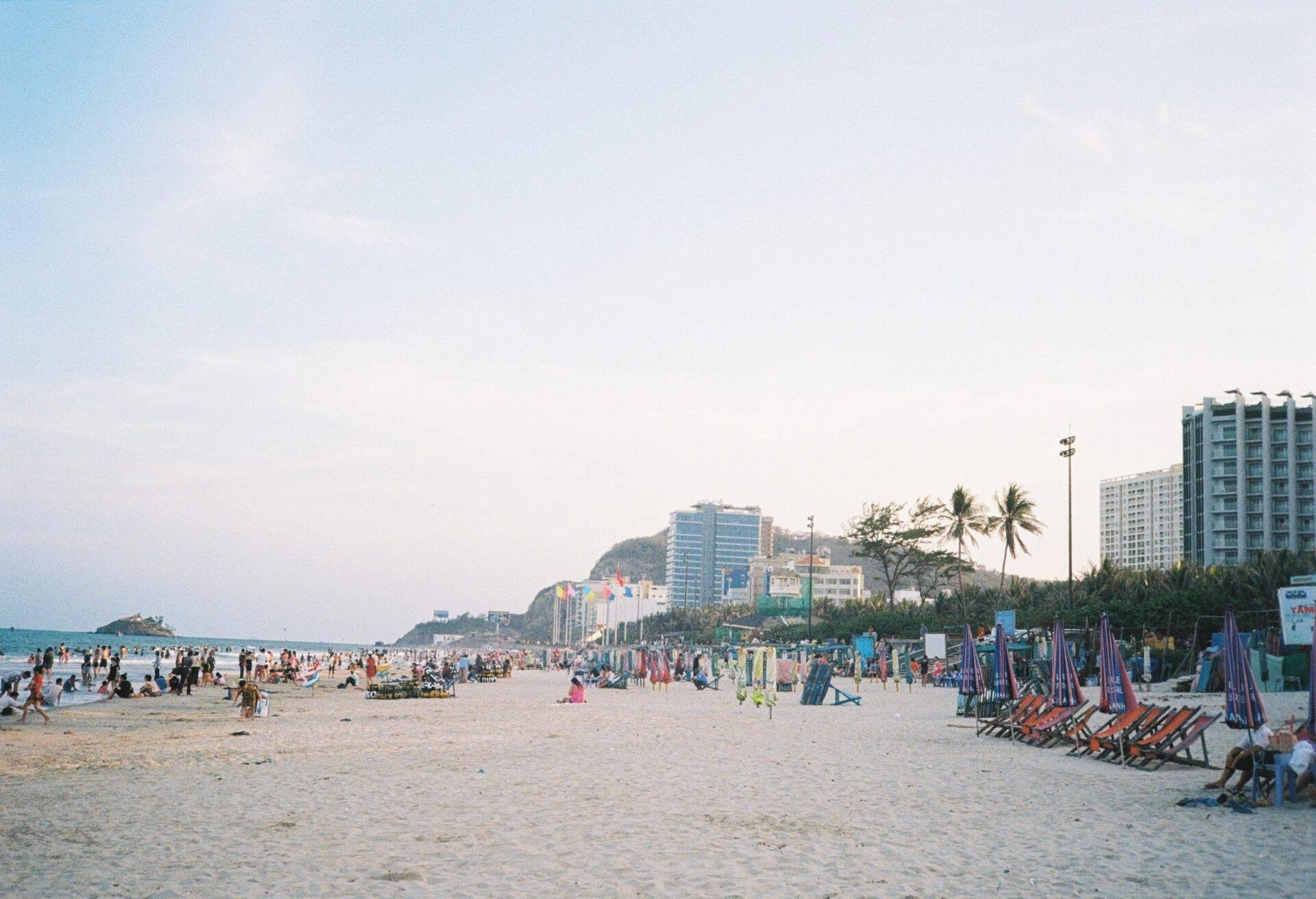 Una playa de arena blanca con tumbonas de colores, edificios modernos y palmeras al fondo