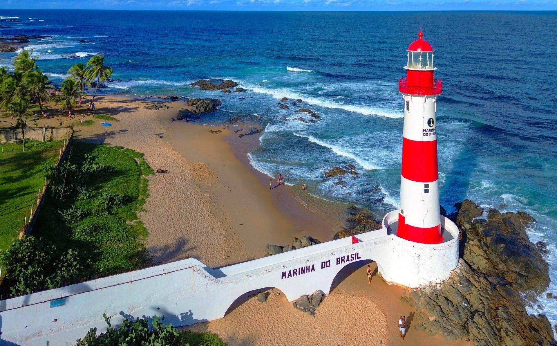 Red and white striped lighthouse on a beachside with palm trees, rocks and blue ocean under a clear sky