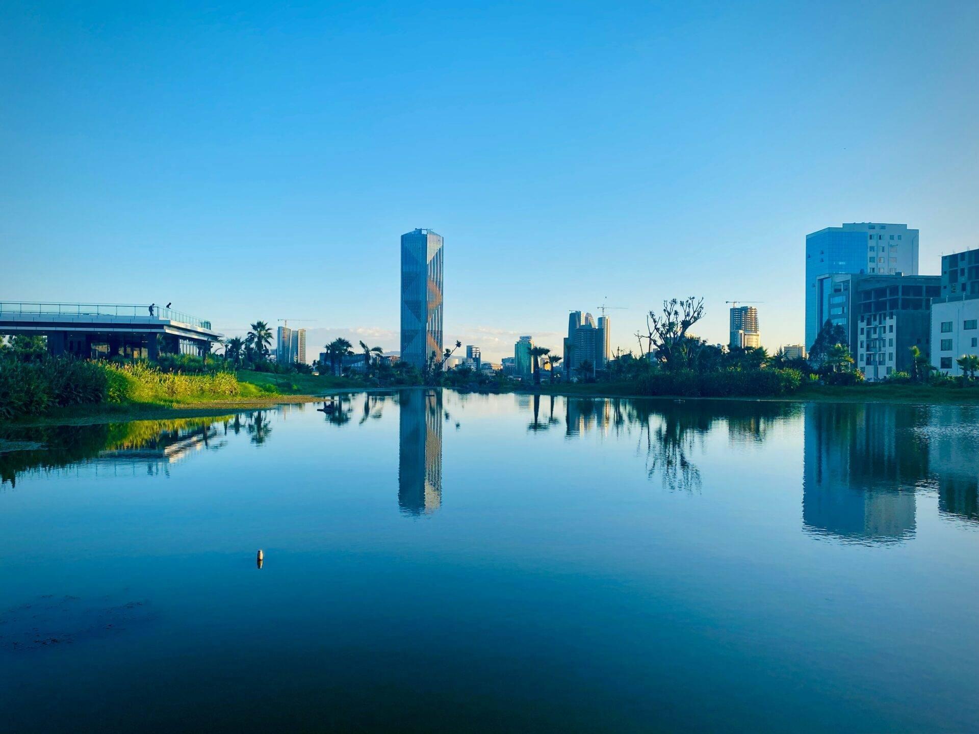 Modern city skyline reflected in the calm waters of a river, with vegetation and a bridge
