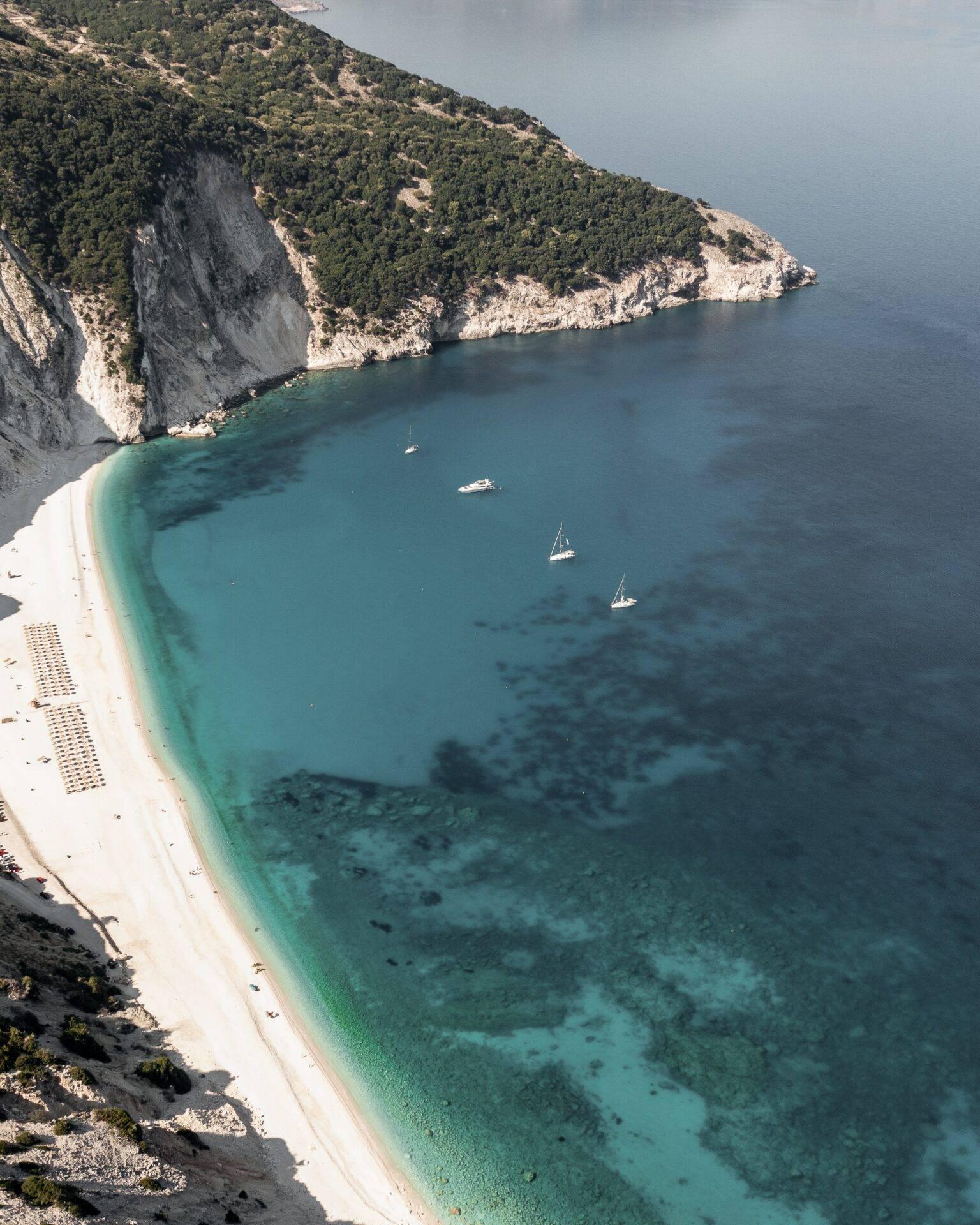 Luftaufnahme eines weißen Sandstrands, gesäumt von Kalksteinklippen, mit türkisfarbenem Wasser und ankernden Segelbooten