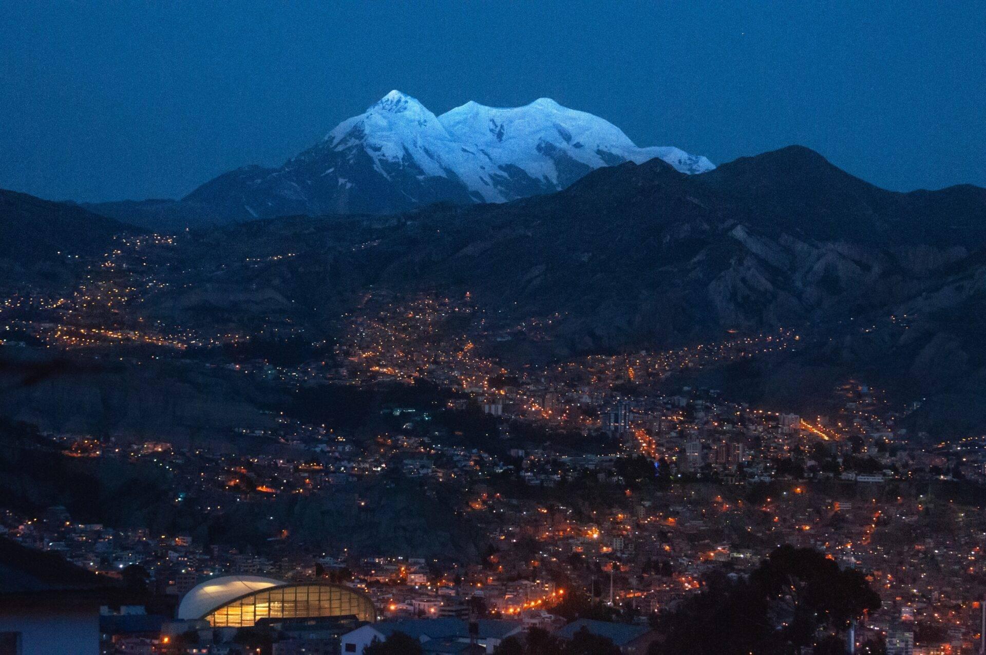 Città illuminata di notte ai piedi di un vulcano innevato sotto un cielo crepuscolare azzurro, con uno stadio moderno in primo piano