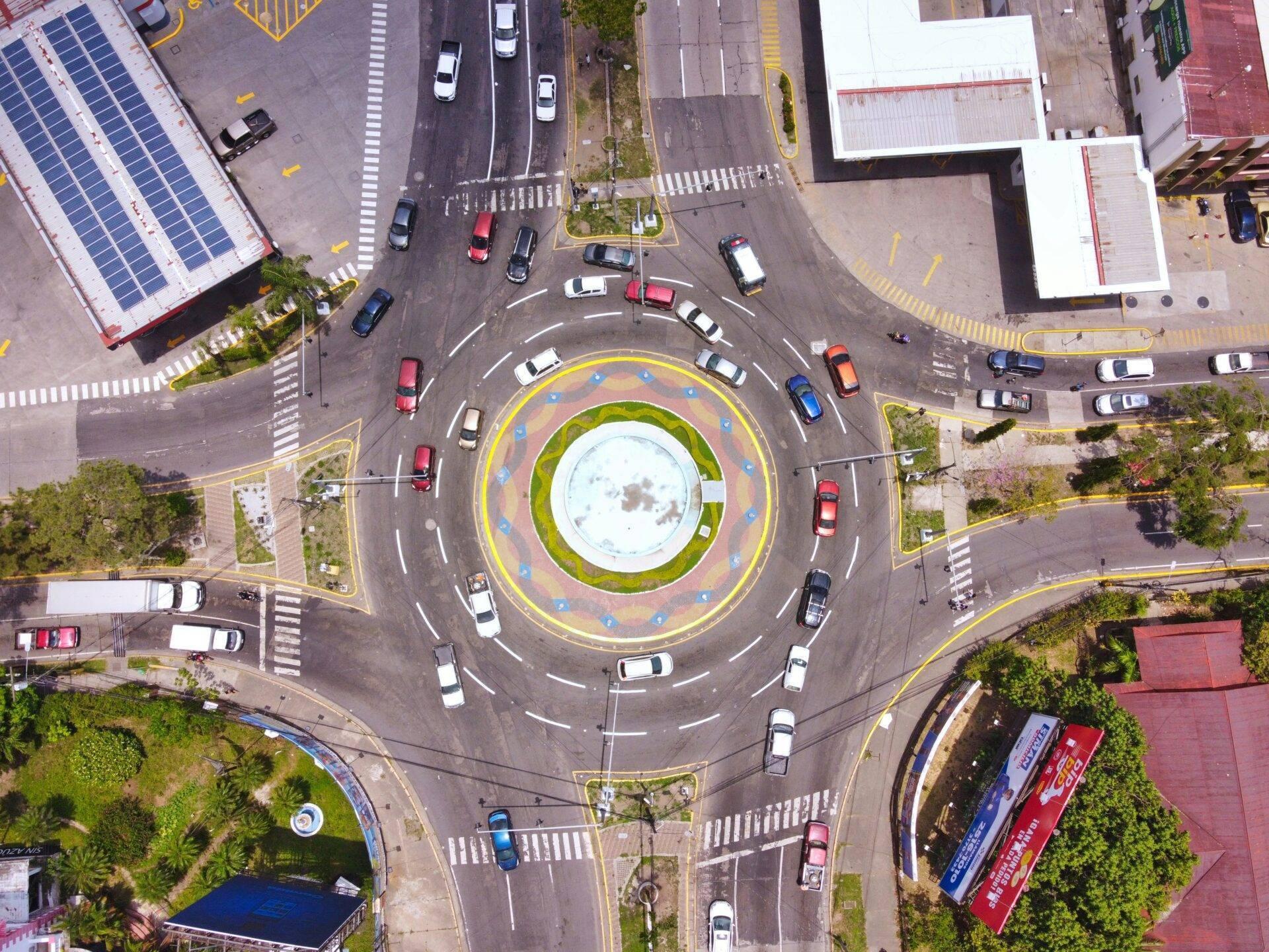 Vista aerea di una rotatoria urbana con fontana centrale, automobili e edifici circostanti con pannelli solari sui tetti
