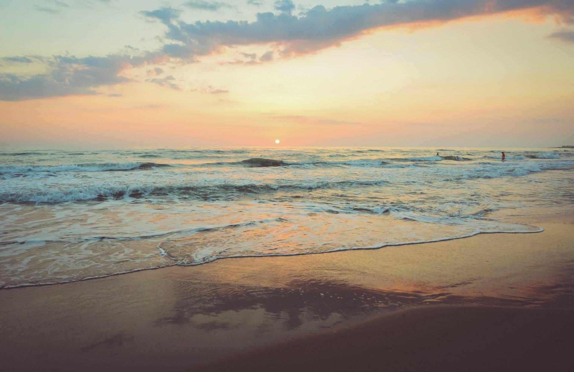Tramonto su una spiaggia di sabbia con onde e qualche bagnante all’orizzonte