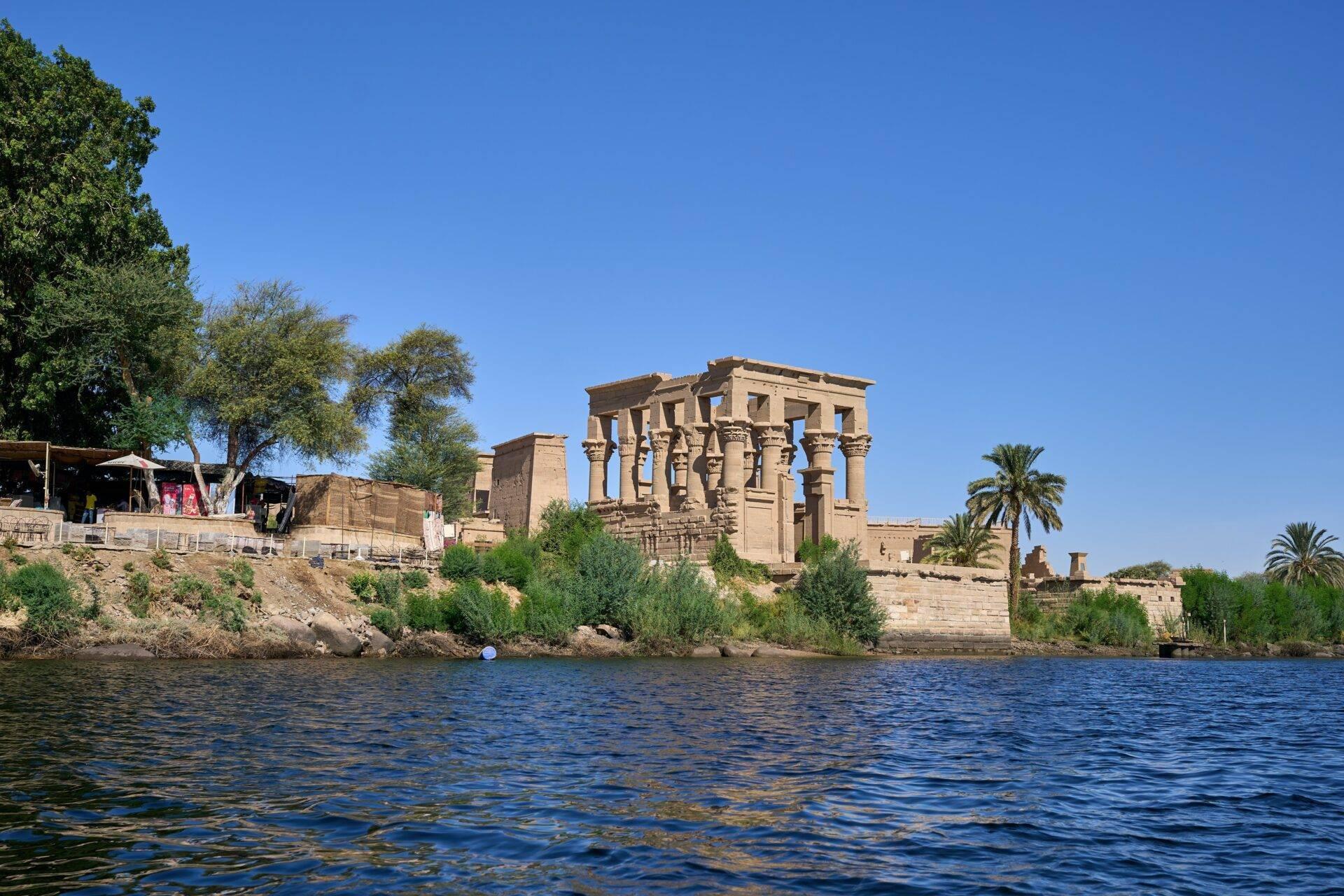 Rovine di un tempio antico sulle rive del Nilo con colonne imponenti e palme sotto un cielo azzurro limpido
