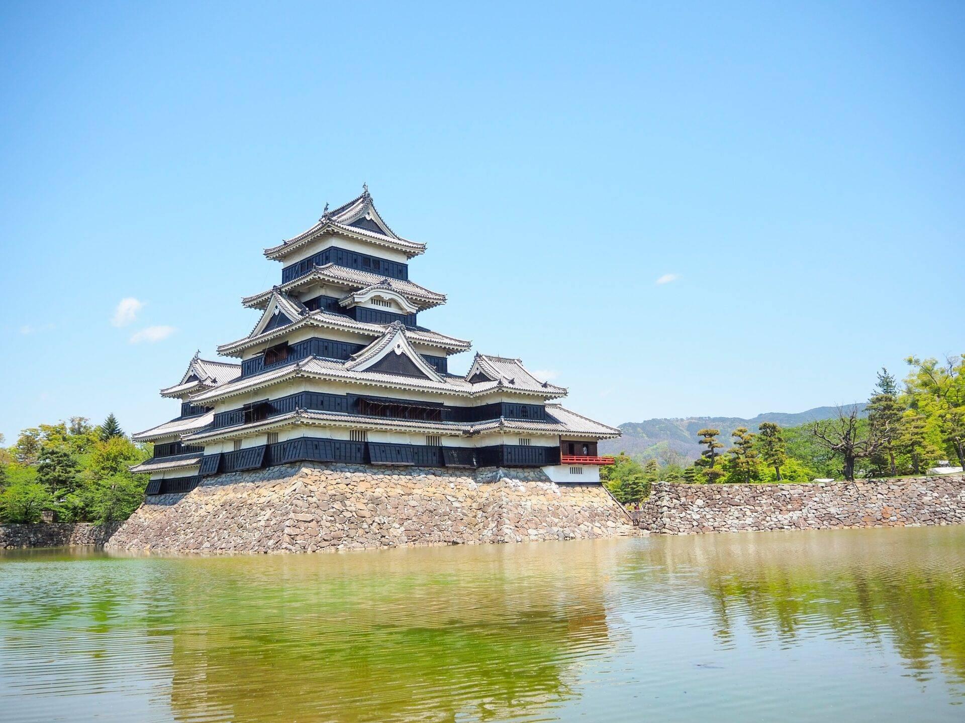 Eine traditionelle japanische Burg mit gestaffelten Dächern überragt einen mit Wasser gefüllten Graben, umgeben von Grün