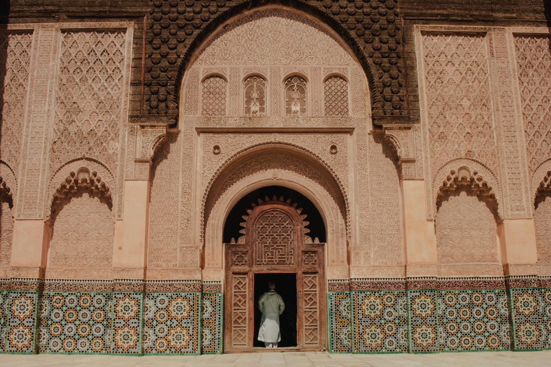 Eine Person in weißer Kleidung vor dem verzierten Eingang einer marokkanischen Moschee mit geometrischen Details und türkisfarbenen Fliesen